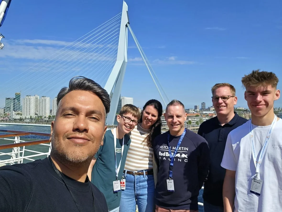 Rotterdam Cruise Port: Our Complete Guide + Insider Tips 2026 (Visited 5 Times Since 2022 by Danny and Peter) 1 Cruising with Danny and Peter, Holland America Line, Nieuw Statendam, Rotterdam, The Netherlands, Voyage Rotterdam Nieuw Statendam Embarkation | A vibrant collage capturing embarkation day on Holland America’s Nieuw Statendam docked at Rotterdam’s Cruise Terminal, with the Erasmus Bridge and Zalmhaventoren in the background under a golden afternoon sky. The ship’s elegant atrium sparkles with a chandelier, while a cozy Oceanview Family Cabin showcases bunk beds and large windows framing the Nieuwe Maas. A family tours the Lido Market, where tables overflow with herb-crusted lamb, chocolate eggs, and colorful desserts for Easter lunch. On deck, two teenagers snap selfies against Rotterdam’s twinkling skyline during sail away, the ship gliding past the illuminated Swan Bridge. In the Ocean Bar, friends toast with cocktails, and the Crow’s Nest lounge offers panoramic views of the river. The scene closes with a cozy cabin moment, a TV glowing with Formula 1 action, hot chocolate mugs on a table, and the gentle sway of the ship at dusk.