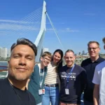 Cruising with Danny and Peter, Holland America Line, Nieuw Statendam, Rotterdam, The Netherlands, Voyage Rotterdam Nieuw Statendam Embarkation | A vibrant collage capturing embarkation day on Holland America’s Nieuw Statendam docked at Rotterdam’s Cruise Terminal, with the Erasmus Bridge and Zalmhaventoren in the background under a golden afternoon sky. The ship’s elegant atrium sparkles with a chandelier, while a cozy Oceanview Family Cabin showcases bunk beds and large windows framing the Nieuwe Maas. A family tours the Lido Market, where tables overflow with herb-crusted lamb, chocolate eggs, and colorful desserts for Easter lunch. On deck, two teenagers snap selfies against Rotterdam’s twinkling skyline during sail away, the ship gliding past the illuminated Swan Bridge. In the Ocean Bar, friends toast with cocktails, and the Crow’s Nest lounge offers panoramic views of the river. The scene closes with a cozy cabin moment, a TV glowing with Formula 1 action, hot chocolate mugs on a table, and the gentle sway of the ship at dusk.