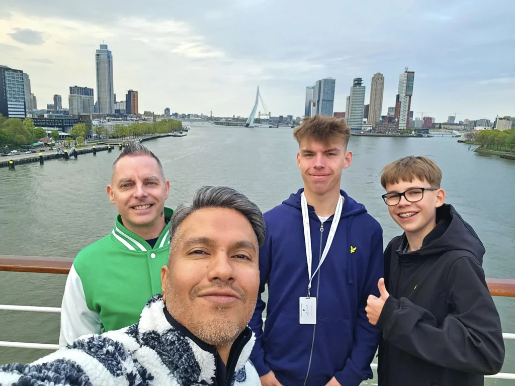 Rotterdam Cruise Port: Our Complete Guide + Insider Tips 2026 (Visited 5 Times Since 2022 by Danny and Peter) 14 Cruising with Danny and Peter, Holland America Line, Nieuw Statendam, Rotterdam, The Netherlands, Voyage Rotterdam Nieuw Statendam Embarkation | A vibrant collage capturing embarkation day on Holland America’s Nieuw Statendam docked at Rotterdam’s Cruise Terminal, with the Erasmus Bridge and Zalmhaventoren in the background under a golden afternoon sky. The ship’s elegant atrium sparkles with a chandelier, while a cozy Oceanview Family Cabin showcases bunk beds and large windows framing the Nieuwe Maas. A family tours the Lido Market, where tables overflow with herb-crusted lamb, chocolate eggs, and colorful desserts for Easter lunch. On deck, two teenagers snap selfies against Rotterdam’s twinkling skyline during sail away, the ship gliding past the illuminated Swan Bridge. In the Ocean Bar, friends toast with cocktails, and the Crow’s Nest lounge offers panoramic views of the river. The scene closes with a cozy cabin moment, a TV glowing with Formula 1 action, hot chocolate mugs on a table, and the gentle sway of the ship at dusk.