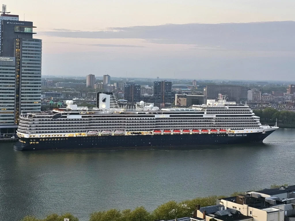 Rotterdam Cruise Port: Our Complete Guide + Insider Tips 2026 (Visited 5 Times Since 2022 by Danny and Peter) 15 Cruising with Danny and Peter, Holland America Line, Nieuw Statendam, Rotterdam, The Netherlands, Voyage Rotterdam Nieuw Statendam Embarkation | A vibrant collage capturing embarkation day on Holland America’s Nieuw Statendam docked at Rotterdam’s Cruise Terminal, with the Erasmus Bridge and Zalmhaventoren in the background under a golden afternoon sky. The ship’s elegant atrium sparkles with a chandelier, while a cozy Oceanview Family Cabin showcases bunk beds and large windows framing the Nieuwe Maas. A family tours the Lido Market, where tables overflow with herb-crusted lamb, chocolate eggs, and colorful desserts for Easter lunch. On deck, two teenagers snap selfies against Rotterdam’s twinkling skyline during sail away, the ship gliding past the illuminated Swan Bridge. In the Ocean Bar, friends toast with cocktails, and the Crow’s Nest lounge offers panoramic views of the river. The scene closes with a cozy cabin moment, a TV glowing with Formula 1 action, hot chocolate mugs on a table, and the gentle sway of the ship at dusk.