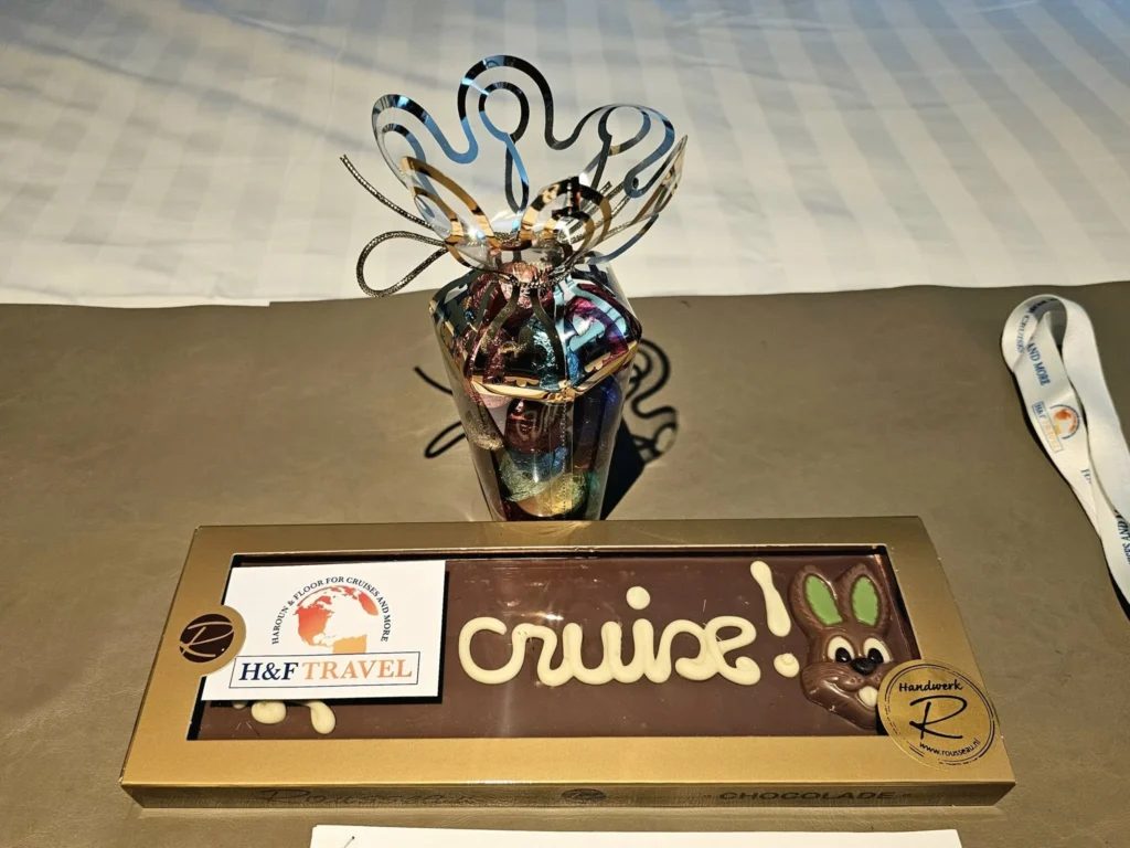 Rotterdam Cruise Port: Our Complete Guide + Insider Tips 2026 (Visited 5 Times Since 2022 by Danny and Peter) 17 Cruising with Danny and Peter, Holland America Line, Nieuw Statendam, Rotterdam, The Netherlands, Voyage Rotterdam Nieuw Statendam Embarkation | A vibrant collage capturing embarkation day on Holland America’s Nieuw Statendam docked at Rotterdam’s Cruise Terminal, with the Erasmus Bridge and Zalmhaventoren in the background under a golden afternoon sky. The ship’s elegant atrium sparkles with a chandelier, while a cozy Oceanview Family Cabin showcases bunk beds and large windows framing the Nieuwe Maas. A family tours the Lido Market, where tables overflow with herb-crusted lamb, chocolate eggs, and colorful desserts for Easter lunch. On deck, two teenagers snap selfies against Rotterdam’s twinkling skyline during sail away, the ship gliding past the illuminated Swan Bridge. In the Ocean Bar, friends toast with cocktails, and the Crow’s Nest lounge offers panoramic views of the river. The scene closes with a cozy cabin moment, a TV glowing with Formula 1 action, hot chocolate mugs on a table, and the gentle sway of the ship at dusk.