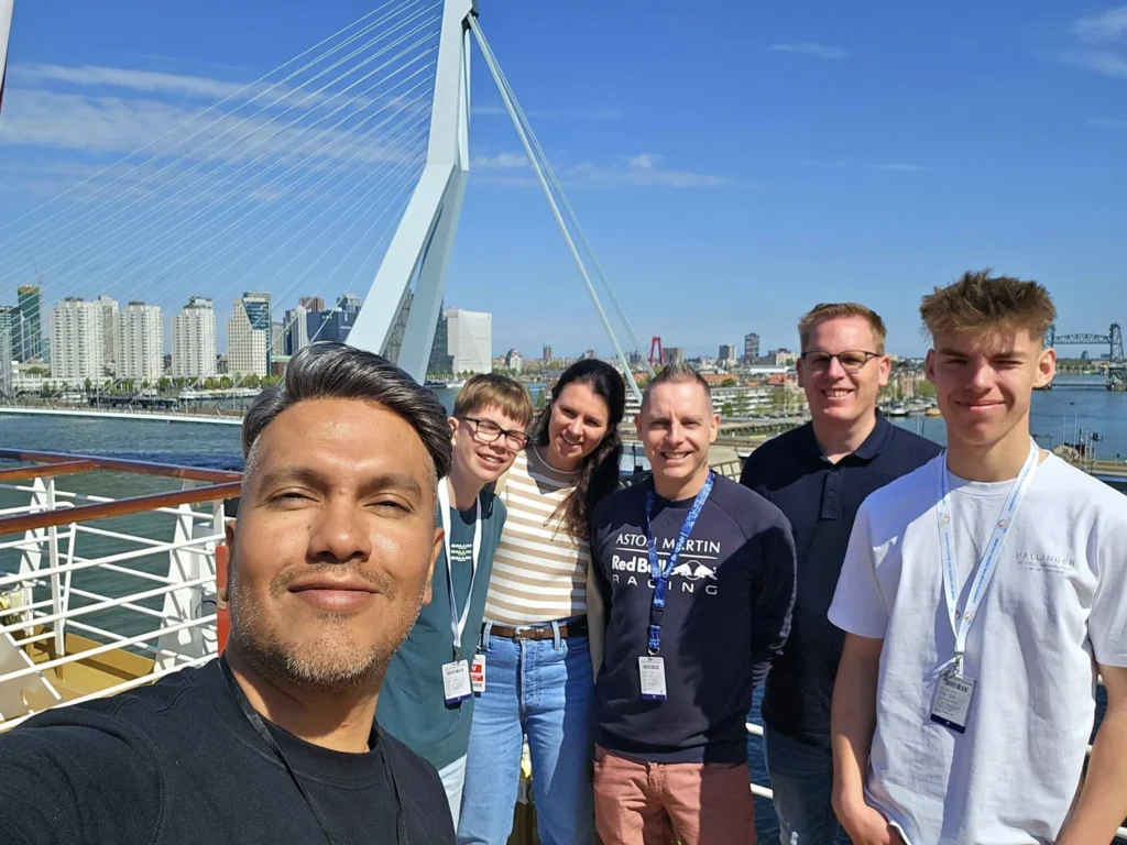 Rotterdam Cruise Port: Our Complete Guide + Insider Tips 2026 (Visited 5 Times Since 2022 by Danny and Peter) 2 Cruising with Danny and Peter, Holland America Line, Nieuw Statendam, Rotterdam, The Netherlands, Voyage Rotterdam Nieuw Statendam Embarkation | A vibrant collage capturing embarkation day on Holland America’s Nieuw Statendam docked at Rotterdam’s Cruise Terminal, with the Erasmus Bridge and Zalmhaventoren in the background under a golden afternoon sky. The ship’s elegant atrium sparkles with a chandelier, while a cozy Oceanview Family Cabin showcases bunk beds and large windows framing the Nieuwe Maas. A family tours the Lido Market, where tables overflow with herb-crusted lamb, chocolate eggs, and colorful desserts for Easter lunch. On deck, two teenagers snap selfies against Rotterdam’s twinkling skyline during sail away, the ship gliding past the illuminated Swan Bridge. In the Ocean Bar, friends toast with cocktails, and the Crow’s Nest lounge offers panoramic views of the river. The scene closes with a cozy cabin moment, a TV glowing with Formula 1 action, hot chocolate mugs on a table, and the gentle sway of the ship at dusk.