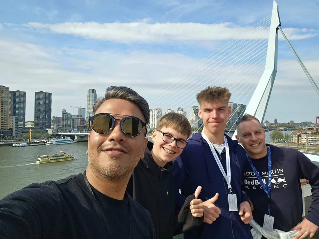 Rotterdam Cruise Port: Our Complete Guide + Insider Tips 2026 (Visited 5 Times Since 2022 by Danny and Peter) 21 Cruising with Danny and Peter, Holland America Line, Nieuw Statendam, Rotterdam, The Netherlands, Voyage Rotterdam Nieuw Statendam Embarkation | A vibrant collage capturing embarkation day on Holland America’s Nieuw Statendam docked at Rotterdam’s Cruise Terminal, with the Erasmus Bridge and Zalmhaventoren in the background under a golden afternoon sky. The ship’s elegant atrium sparkles with a chandelier, while a cozy Oceanview Family Cabin showcases bunk beds and large windows framing the Nieuwe Maas. A family tours the Lido Market, where tables overflow with herb-crusted lamb, chocolate eggs, and colorful desserts for Easter lunch. On deck, two teenagers snap selfies against Rotterdam’s twinkling skyline during sail away, the ship gliding past the illuminated Swan Bridge. In the Ocean Bar, friends toast with cocktails, and the Crow’s Nest lounge offers panoramic views of the river. The scene closes with a cozy cabin moment, a TV glowing with Formula 1 action, hot chocolate mugs on a table, and the gentle sway of the ship at dusk.