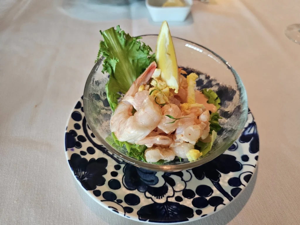 Rotterdam Cruise Port: Our Complete Guide + Insider Tips 2026 (Visited 5 Times Since 2022 by Danny and Peter) 22 Cruising with Danny and Peter, Holland America Line, Nieuw Statendam, Rotterdam, The Netherlands, Voyage Rotterdam Nieuw Statendam Embarkation | A vibrant collage capturing embarkation day on Holland America’s Nieuw Statendam docked at Rotterdam’s Cruise Terminal, with the Erasmus Bridge and Zalmhaventoren in the background under a golden afternoon sky. The ship’s elegant atrium sparkles with a chandelier, while a cozy Oceanview Family Cabin showcases bunk beds and large windows framing the Nieuwe Maas. A family tours the Lido Market, where tables overflow with herb-crusted lamb, chocolate eggs, and colorful desserts for Easter lunch. On deck, two teenagers snap selfies against Rotterdam’s twinkling skyline during sail away, the ship gliding past the illuminated Swan Bridge. In the Ocean Bar, friends toast with cocktails, and the Crow’s Nest lounge offers panoramic views of the river. The scene closes with a cozy cabin moment, a TV glowing with Formula 1 action, hot chocolate mugs on a table, and the gentle sway of the ship at dusk.