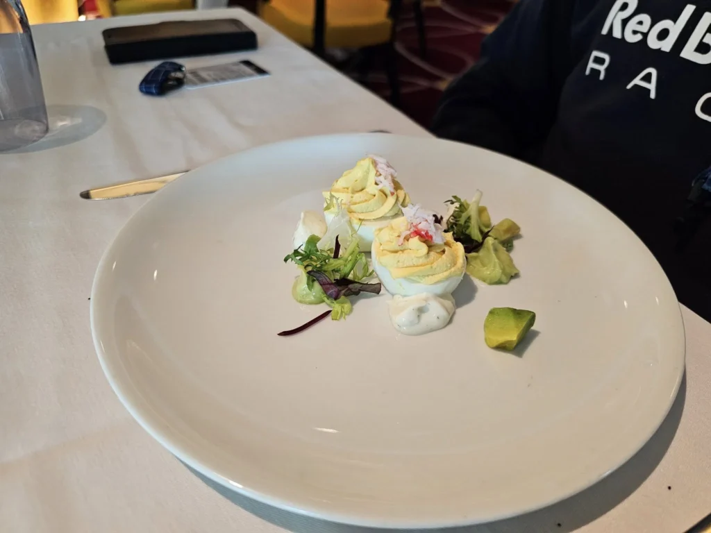 Rotterdam Cruise Port: Our Complete Guide + Insider Tips 2026 (Visited 5 Times Since 2022 by Danny and Peter) 23 Cruising with Danny and Peter, Holland America Line, Nieuw Statendam, Rotterdam, The Netherlands, Voyage Rotterdam Nieuw Statendam Embarkation | A vibrant collage capturing embarkation day on Holland America’s Nieuw Statendam docked at Rotterdam’s Cruise Terminal, with the Erasmus Bridge and Zalmhaventoren in the background under a golden afternoon sky. The ship’s elegant atrium sparkles with a chandelier, while a cozy Oceanview Family Cabin showcases bunk beds and large windows framing the Nieuwe Maas. A family tours the Lido Market, where tables overflow with herb-crusted lamb, chocolate eggs, and colorful desserts for Easter lunch. On deck, two teenagers snap selfies against Rotterdam’s twinkling skyline during sail away, the ship gliding past the illuminated Swan Bridge. In the Ocean Bar, friends toast with cocktails, and the Crow’s Nest lounge offers panoramic views of the river. The scene closes with a cozy cabin moment, a TV glowing with Formula 1 action, hot chocolate mugs on a table, and the gentle sway of the ship at dusk.