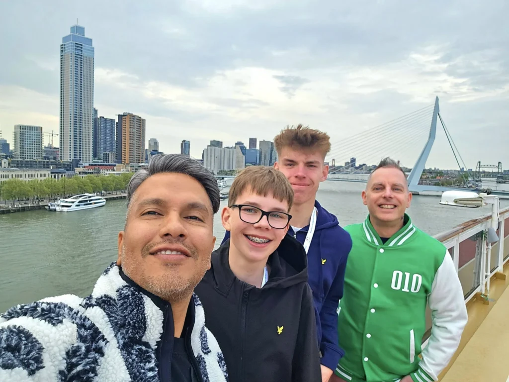 Rotterdam Cruise Port: Our Complete Guide + Insider Tips 2026 (Visited 5 Times Since 2022 by Danny and Peter) 6 Cruising with Danny and Peter, Holland America Line, Nieuw Statendam, Rotterdam, The Netherlands, Voyage Rotterdam Nieuw Statendam Embarkation | A vibrant collage capturing embarkation day on Holland America’s Nieuw Statendam docked at Rotterdam’s Cruise Terminal, with the Erasmus Bridge and Zalmhaventoren in the background under a golden afternoon sky. The ship’s elegant atrium sparkles with a chandelier, while a cozy Oceanview Family Cabin showcases bunk beds and large windows framing the Nieuwe Maas. A family tours the Lido Market, where tables overflow with herb-crusted lamb, chocolate eggs, and colorful desserts for Easter lunch. On deck, two teenagers snap selfies against Rotterdam’s twinkling skyline during sail away, the ship gliding past the illuminated Swan Bridge. In the Ocean Bar, friends toast with cocktails, and the Crow’s Nest lounge offers panoramic views of the river. The scene closes with a cozy cabin moment, a TV glowing with Formula 1 action, hot chocolate mugs on a table, and the gentle sway of the ship at dusk.