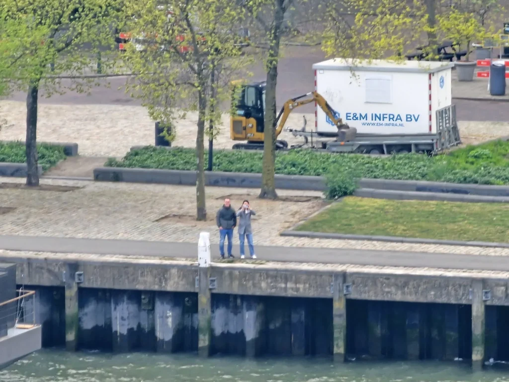 Rotterdam Cruise Port: Our Complete Guide + Insider Tips 2026 (Visited 5 Times Since 2022 by Danny and Peter) 11 Cruising with Danny and Peter, Holland America Line, Nieuw Statendam, Rotterdam, The Netherlands, Voyage Rotterdam Nieuw Statendam Embarkation | A vibrant collage capturing embarkation day on Holland America’s Nieuw Statendam docked at Rotterdam’s Cruise Terminal, with the Erasmus Bridge and Zalmhaventoren in the background under a golden afternoon sky. The ship’s elegant atrium sparkles with a chandelier, while a cozy Oceanview Family Cabin showcases bunk beds and large windows framing the Nieuwe Maas. A family tours the Lido Market, where tables overflow with herb-crusted lamb, chocolate eggs, and colorful desserts for Easter lunch. On deck, two teenagers snap selfies against Rotterdam’s twinkling skyline during sail away, the ship gliding past the illuminated Swan Bridge. In the Ocean Bar, friends toast with cocktails, and the Crow’s Nest lounge offers panoramic views of the river. The scene closes with a cozy cabin moment, a TV glowing with Formula 1 action, hot chocolate mugs on a table, and the gentle sway of the ship at dusk.