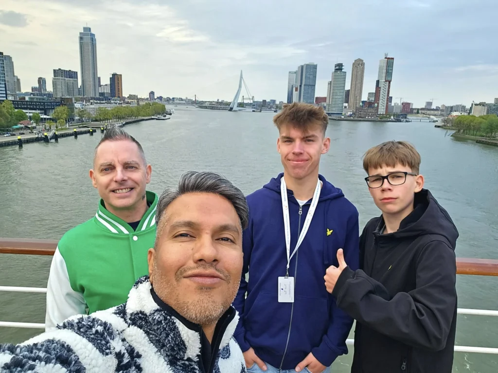 Rotterdam Cruise Port: Our Complete Guide + Insider Tips 2026 (Visited 5 Times Since 2022 by Danny and Peter) 29 Cruising with Danny and Peter, Holland America Line, Nieuw Statendam, Rotterdam, The Netherlands, Voyage Rotterdam Nieuw Statendam Embarkation | A vibrant collage capturing embarkation day on Holland America’s Nieuw Statendam docked at Rotterdam’s Cruise Terminal, with the Erasmus Bridge and Zalmhaventoren in the background under a golden afternoon sky. The ship’s elegant atrium sparkles with a chandelier, while a cozy Oceanview Family Cabin showcases bunk beds and large windows framing the Nieuwe Maas. A family tours the Lido Market, where tables overflow with herb-crusted lamb, chocolate eggs, and colorful desserts for Easter lunch. On deck, two teenagers snap selfies against Rotterdam’s twinkling skyline during sail away, the ship gliding past the illuminated Swan Bridge. In the Ocean Bar, friends toast with cocktails, and the Crow’s Nest lounge offers panoramic views of the river. The scene closes with a cozy cabin moment, a TV glowing with Formula 1 action, hot chocolate mugs on a table, and the gentle sway of the ship at dusk.