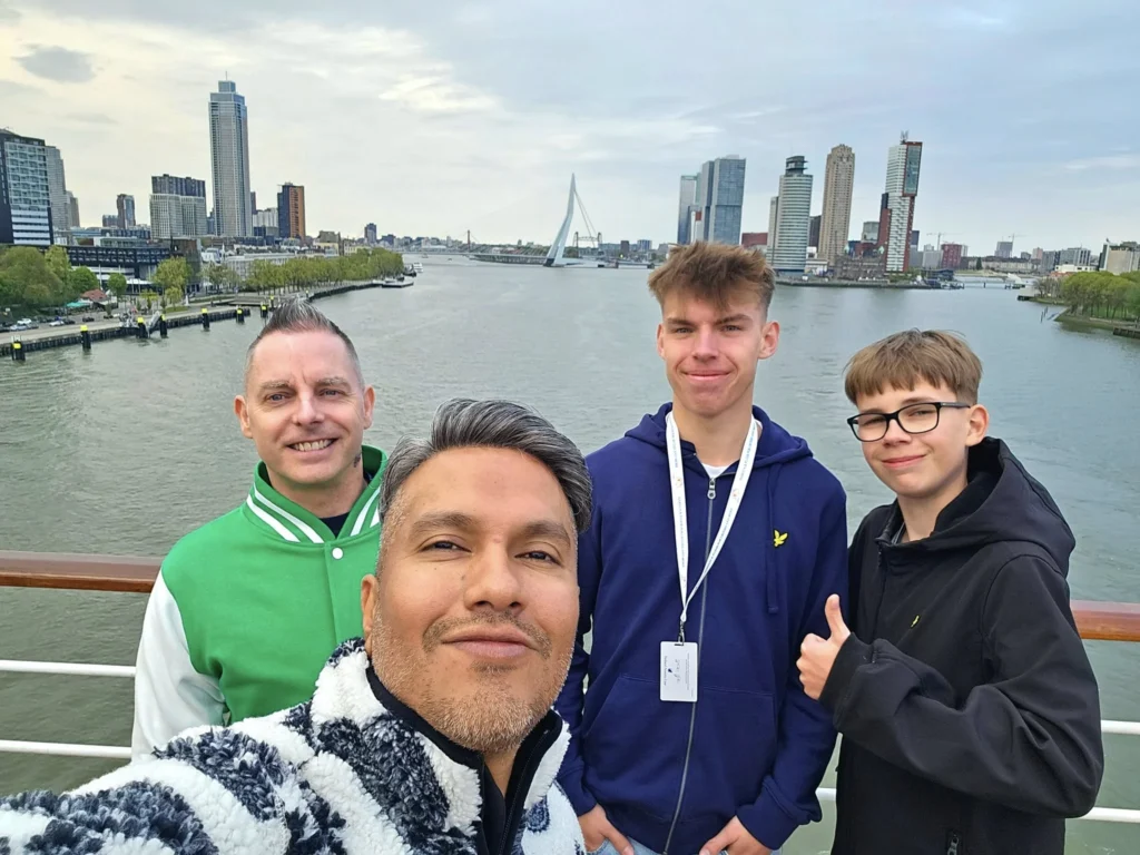 Rotterdam Cruise Port: Our Complete Guide + Insider Tips 2026 (Visited 5 Times Since 2022 by Danny and Peter) 30 Cruising with Danny and Peter, Holland America Line, Nieuw Statendam, Rotterdam, The Netherlands, Voyage Rotterdam Nieuw Statendam Embarkation | A vibrant collage capturing embarkation day on Holland America’s Nieuw Statendam docked at Rotterdam’s Cruise Terminal, with the Erasmus Bridge and Zalmhaventoren in the background under a golden afternoon sky. The ship’s elegant atrium sparkles with a chandelier, while a cozy Oceanview Family Cabin showcases bunk beds and large windows framing the Nieuwe Maas. A family tours the Lido Market, where tables overflow with herb-crusted lamb, chocolate eggs, and colorful desserts for Easter lunch. On deck, two teenagers snap selfies against Rotterdam’s twinkling skyline during sail away, the ship gliding past the illuminated Swan Bridge. In the Ocean Bar, friends toast with cocktails, and the Crow’s Nest lounge offers panoramic views of the river. The scene closes with a cozy cabin moment, a TV glowing with Formula 1 action, hot chocolate mugs on a table, and the gentle sway of the ship at dusk.