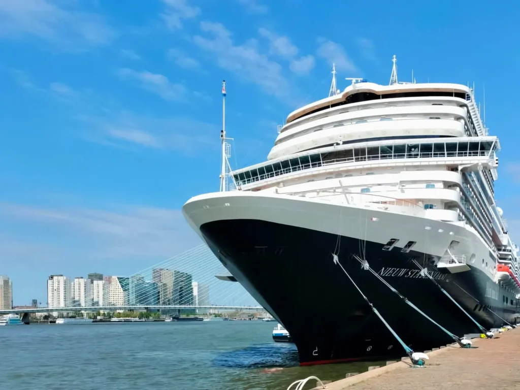 Rotterdam Cruise Port: Our Complete Guide + Insider Tips 2026 (Visited 5 Times Since 2022 by Danny and Peter) 31 Cruising with Danny and Peter, Holland America Line, Nieuw Statendam, Rotterdam, The Netherlands, Voyage Rotterdam Nieuw Statendam Embarkation | A vibrant collage capturing embarkation day on Holland America’s Nieuw Statendam docked at Rotterdam’s Cruise Terminal, with the Erasmus Bridge and Zalmhaventoren in the background under a golden afternoon sky. The ship’s elegant atrium sparkles with a chandelier, while a cozy Oceanview Family Cabin showcases bunk beds and large windows framing the Nieuwe Maas. A family tours the Lido Market, where tables overflow with herb-crusted lamb, chocolate eggs, and colorful desserts for Easter lunch. On deck, two teenagers snap selfies against Rotterdam’s twinkling skyline during sail away, the ship gliding past the illuminated Swan Bridge. In the Ocean Bar, friends toast with cocktails, and the Crow’s Nest lounge offers panoramic views of the river. The scene closes with a cozy cabin moment, a TV glowing with Formula 1 action, hot chocolate mugs on a table, and the gentle sway of the ship at dusk.