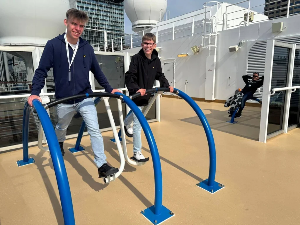 Rotterdam Cruise Port: Our Complete Guide + Insider Tips 2026 (Visited 5 Times Since 2022 by Danny and Peter) 32 Cruising with Danny and Peter, Holland America Line, Nieuw Statendam, Rotterdam, The Netherlands, Voyage Rotterdam Nieuw Statendam Embarkation | A vibrant collage capturing embarkation day on Holland America’s Nieuw Statendam docked at Rotterdam’s Cruise Terminal, with the Erasmus Bridge and Zalmhaventoren in the background under a golden afternoon sky. The ship’s elegant atrium sparkles with a chandelier, while a cozy Oceanview Family Cabin showcases bunk beds and large windows framing the Nieuwe Maas. A family tours the Lido Market, where tables overflow with herb-crusted lamb, chocolate eggs, and colorful desserts for Easter lunch. On deck, two teenagers snap selfies against Rotterdam’s twinkling skyline during sail away, the ship gliding past the illuminated Swan Bridge. In the Ocean Bar, friends toast with cocktails, and the Crow’s Nest lounge offers panoramic views of the river. The scene closes with a cozy cabin moment, a TV glowing with Formula 1 action, hot chocolate mugs on a table, and the gentle sway of the ship at dusk.