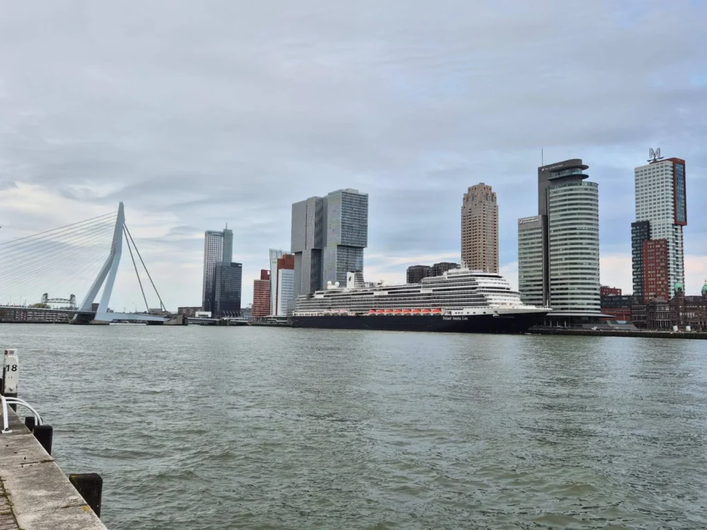 Rotterdam Cruise Port: Our Complete Guide + Insider Tips 2026 (Visited 5 Times Since 2022 by Danny and Peter) 12 Cruising with Danny and Peter, Holland America Line, Nieuw Statendam, Rotterdam, The Netherlands, Voyage Rotterdam Nieuw Statendam Embarkation | A vibrant collage capturing embarkation day on Holland America’s Nieuw Statendam docked at Rotterdam’s Cruise Terminal, with the Erasmus Bridge and Zalmhaventoren in the background under a golden afternoon sky. The ship’s elegant atrium sparkles with a chandelier, while a cozy Oceanview Family Cabin showcases bunk beds and large windows framing the Nieuwe Maas. A family tours the Lido Market, where tables overflow with herb-crusted lamb, chocolate eggs, and colorful desserts for Easter lunch. On deck, two teenagers snap selfies against Rotterdam’s twinkling skyline during sail away, the ship gliding past the illuminated Swan Bridge. In the Ocean Bar, friends toast with cocktails, and the Crow’s Nest lounge offers panoramic views of the river. The scene closes with a cozy cabin moment, a TV glowing with Formula 1 action, hot chocolate mugs on a table, and the gentle sway of the ship at dusk.