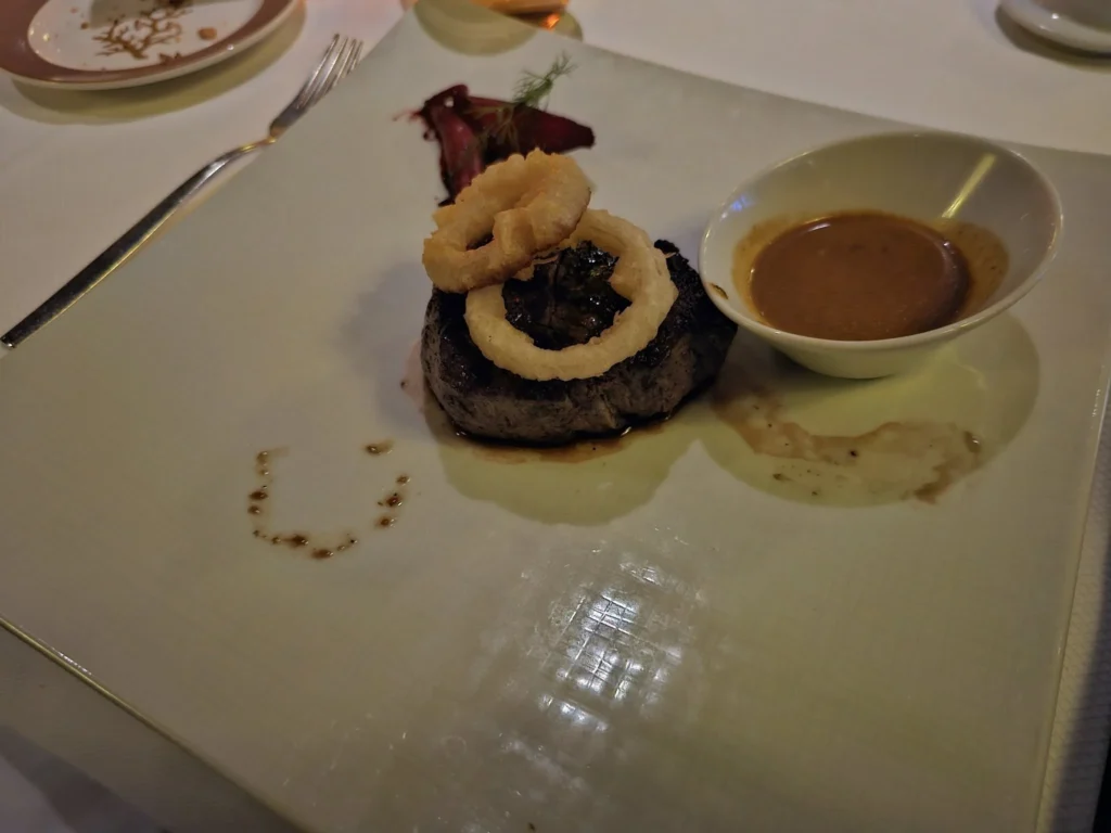 A Voyage Through Time and Tide: Exploring the English Channel Aboard Holland America Line’s Nieuw Statendam 28 Cruising with Danny and Peter, Holland America Line, Nieuw Statendam, English Channel, Sea Day, English Channel Nieuw Statendam Sea Day Experience | A luxurious scene aboard Holland America Line’s Nieuw Statendam cruising the English Channel, featuring the Main Dining Room with elegantly set tables, gourmet dishes like eggs Benedict, and large windows revealing gray-green waves; the intimate Pinnacle Grill with plush seating and a plated beef tenderloin with lobster dumplings; the World Stage theater’s circular stage and vibrant 270-degree LED screen displaying Casablanca’s Hassan II Mosque; and the ship’s sleek white hull sailing smoothly under an overcast sky, embodying modern cruising elegance.