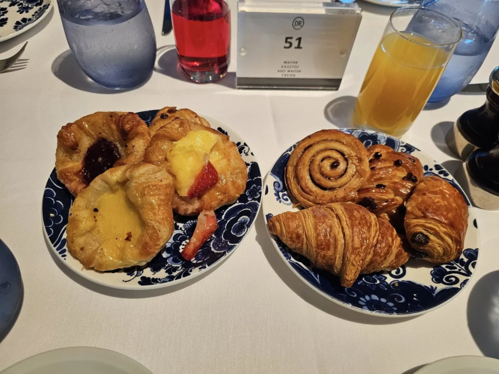 Cruising with Danny and Peter, Holland America Line, Nieuw Statendam, Bay Of Biscay History Sea Day | A scenic view of the Bay of Biscay’s deep blue waters under a partly cloudy sky, with gentle waves reflecting sunlight, alongside the elegant Dining Room of Holland America’s Nieuw Statendam, featuring white tablecloths, polished silverware, and a plate of lobster Benedict with hollandaise sauce. The World Stage theater showcases a vibrant LED screen displaying Agadir’s bustling souk with colorful spices and crafts, while the Lido Pool area glows at night with twinkling lights, where guests mingle with champagne during the Mariners Reception. The intimate Tamarind restaurant, with warm wood decor, presents a beautifully plated Penang red curry with duck, paired with a sake flight.