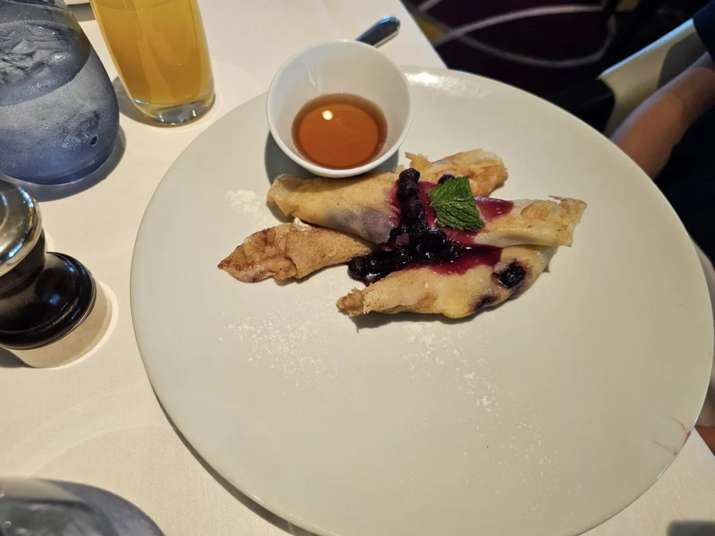 Cruising with Danny and Peter, Holland America Line, Nieuw Statendam, Bay Of Biscay History Sea Day | A scenic view of the Bay of Biscay’s deep blue waters under a partly cloudy sky, with gentle waves reflecting sunlight, alongside the elegant Dining Room of Holland America’s Nieuw Statendam, featuring white tablecloths, polished silverware, and a plate of lobster Benedict with hollandaise sauce. The World Stage theater showcases a vibrant LED screen displaying Agadir’s bustling souk with colorful spices and crafts, while the Lido Pool area glows at night with twinkling lights, where guests mingle with champagne during the Mariners Reception. The intimate Tamarind restaurant, with warm wood decor, presents a beautifully plated Penang red curry with duck, paired with a sake flight.