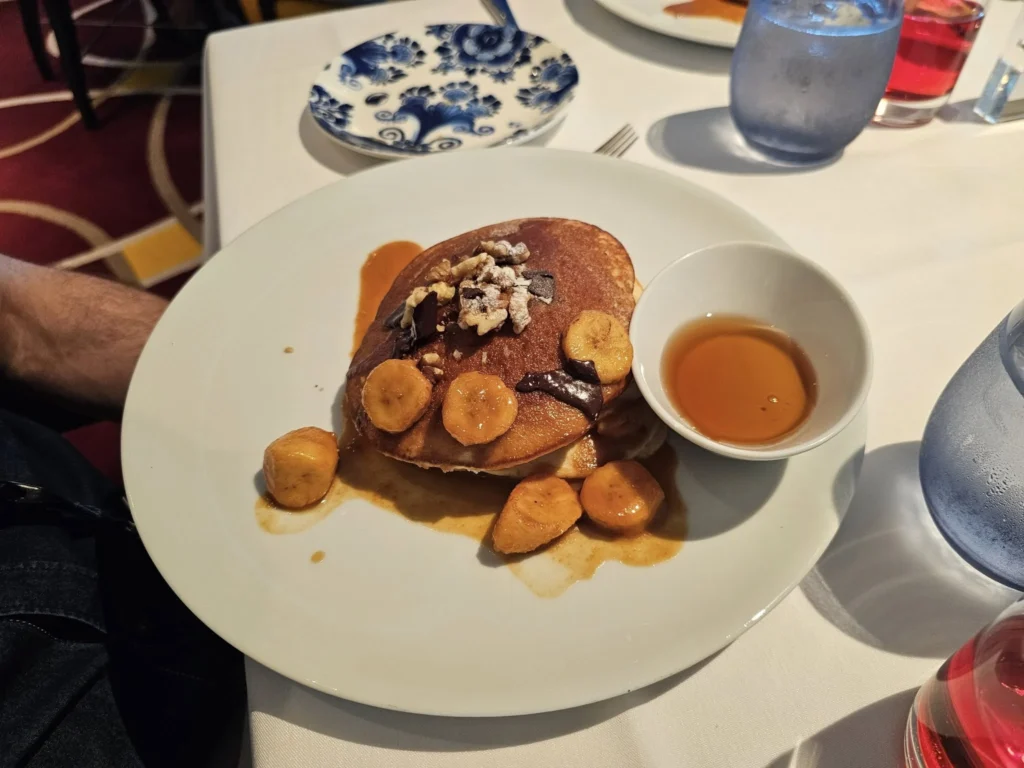 Cruising with Danny and Peter, Holland America Line, Nieuw Statendam, Bay Of Biscay History Sea Day | A scenic view of the Bay of Biscay’s deep blue waters under a partly cloudy sky, with gentle waves reflecting sunlight, alongside the elegant Dining Room of Holland America’s Nieuw Statendam, featuring white tablecloths, polished silverware, and a plate of lobster Benedict with hollandaise sauce. The World Stage theater showcases a vibrant LED screen displaying Agadir’s bustling souk with colorful spices and crafts, while the Lido Pool area glows at night with twinkling lights, where guests mingle with champagne during the Mariners Reception. The intimate Tamarind restaurant, with warm wood decor, presents a beautifully plated Penang red curry with duck, paired with a sake flight.