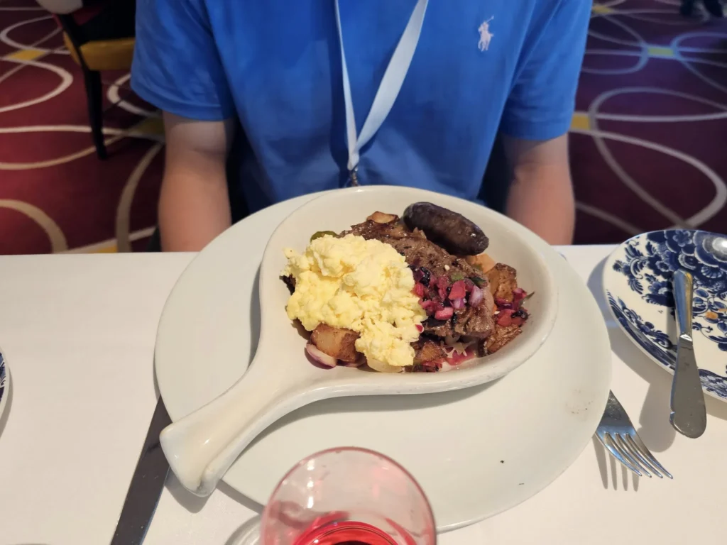 Cruising with Danny and Peter, Holland America Line, Nieuw Statendam, Bay Of Biscay History Sea Day | A scenic view of the Bay of Biscay’s deep blue waters under a partly cloudy sky, with gentle waves reflecting sunlight, alongside the elegant Dining Room of Holland America’s Nieuw Statendam, featuring white tablecloths, polished silverware, and a plate of lobster Benedict with hollandaise sauce. The World Stage theater showcases a vibrant LED screen displaying Agadir’s bustling souk with colorful spices and crafts, while the Lido Pool area glows at night with twinkling lights, where guests mingle with champagne during the Mariners Reception. The intimate Tamarind restaurant, with warm wood decor, presents a beautifully plated Penang red curry with duck, paired with a sake flight.