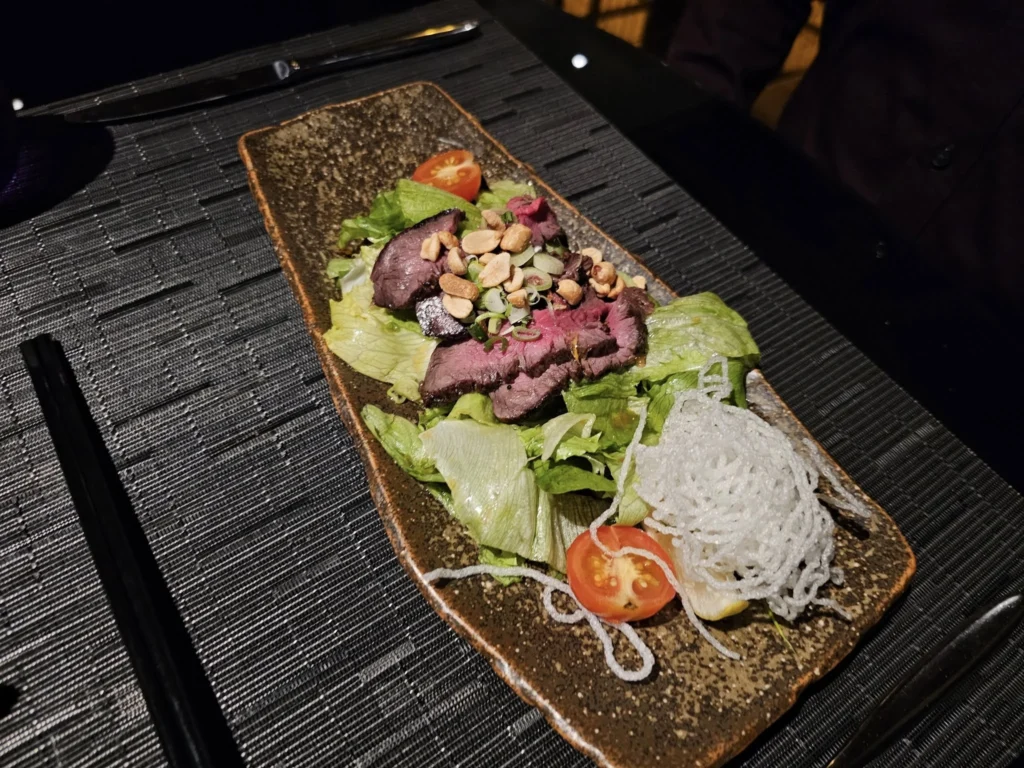 Cruising with Danny and Peter, Holland America Line, Nieuw Statendam, Bay Of Biscay History Sea Day | A scenic view of the Bay of Biscay’s deep blue waters under a partly cloudy sky, with gentle waves reflecting sunlight, alongside the elegant Dining Room of Holland America’s Nieuw Statendam, featuring white tablecloths, polished silverware, and a plate of lobster Benedict with hollandaise sauce. The World Stage theater showcases a vibrant LED screen displaying Agadir’s bustling souk with colorful spices and crafts, while the Lido Pool area glows at night with twinkling lights, where guests mingle with champagne during the Mariners Reception. The intimate Tamarind restaurant, with warm wood decor, presents a beautifully plated Penang red curry with duck, paired with a sake flight.