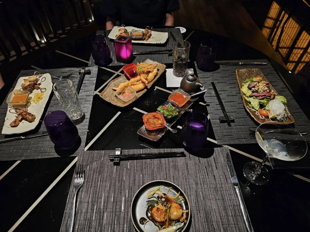 Cruising with Danny and Peter, Holland America Line, Nieuw Statendam, Bay Of Biscay History Sea Day | A scenic view of the Bay of Biscay’s deep blue waters under a partly cloudy sky, with gentle waves reflecting sunlight, alongside the elegant Dining Room of Holland America’s Nieuw Statendam, featuring white tablecloths, polished silverware, and a plate of lobster Benedict with hollandaise sauce. The World Stage theater showcases a vibrant LED screen displaying Agadir’s bustling souk with colorful spices and crafts, while the Lido Pool area glows at night with twinkling lights, where guests mingle with champagne during the Mariners Reception. The intimate Tamarind restaurant, with warm wood decor, presents a beautifully plated Penang red curry with duck, paired with a sake flight.