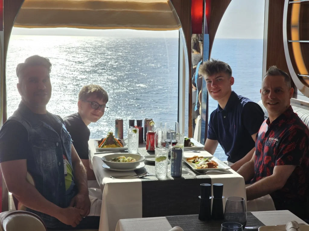 Cruising with Danny and Peter, Holland America Line, Nieuw Statendam, Atlantic Ocean Enchantment Journey Sea Day | A composite image capturing the essence of a luxurious sea day aboard Holland America Line’s Nieuw Statendam cruising the Atlantic Ocean. The scene includes a panoramic view of the vast, shimmering blue Atlantic under a clear sky, with gentle waves and distant seabirds soaring. The elegant interior of the Nieuw Statendam’s Lido Market buffet is shown, featuring a vibrant display of chocolates and cheeses on a table, surrounded by modern decor and large windows framing the ocean. A cozy, warmly lit dining setup at Canaletto, the ship’s Italian specialty restaurant, showcases a beautifully plated osso buco dish with saffron risotto, accompanied by a glass of Chianti. The ship’s sleek exterior glides through the water, its white hull contrasting the deep ocean, with the Mid-Atlantic Ridge subtly illustrated beneath the surface to hint at the ocean’s geological history. A group of passengers enjoys a lively trivia game in the Ocean Bar, with a mysterious camera crew discreetly filming in the background, adding an air of intrigue. The final touch includes a glimpse of a plush stateroom with a large window revealing the moonlit Atlantic, evoking tranquility and anticipation for Agadir, Morocco.