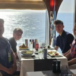 Cruising with Danny and Peter, Holland America Line, Nieuw Statendam, Atlantic Ocean Enchantment Journey Sea Day | A composite image capturing the essence of a luxurious sea day aboard Holland America Line’s Nieuw Statendam cruising the Atlantic Ocean. The scene includes a panoramic view of the vast, shimmering blue Atlantic under a clear sky, with gentle waves and distant seabirds soaring. The elegant interior of the Nieuw Statendam’s Lido Market buffet is shown, featuring a vibrant display of chocolates and cheeses on a table, surrounded by modern decor and large windows framing the ocean. A cozy, warmly lit dining setup at Canaletto, the ship’s Italian specialty restaurant, showcases a beautifully plated osso buco dish with saffron risotto, accompanied by a glass of Chianti. The ship’s sleek exterior glides through the water, its white hull contrasting the deep ocean, with the Mid-Atlantic Ridge subtly illustrated beneath the surface to hint at the ocean’s geological history. A group of passengers enjoys a lively trivia game in the Ocean Bar, with a mysterious camera crew discreetly filming in the background, adding an air of intrigue. The final touch includes a glimpse of a plush stateroom with a large window revealing the moonlit Atlantic, evoking tranquility and anticipation for Agadir, Morocco.