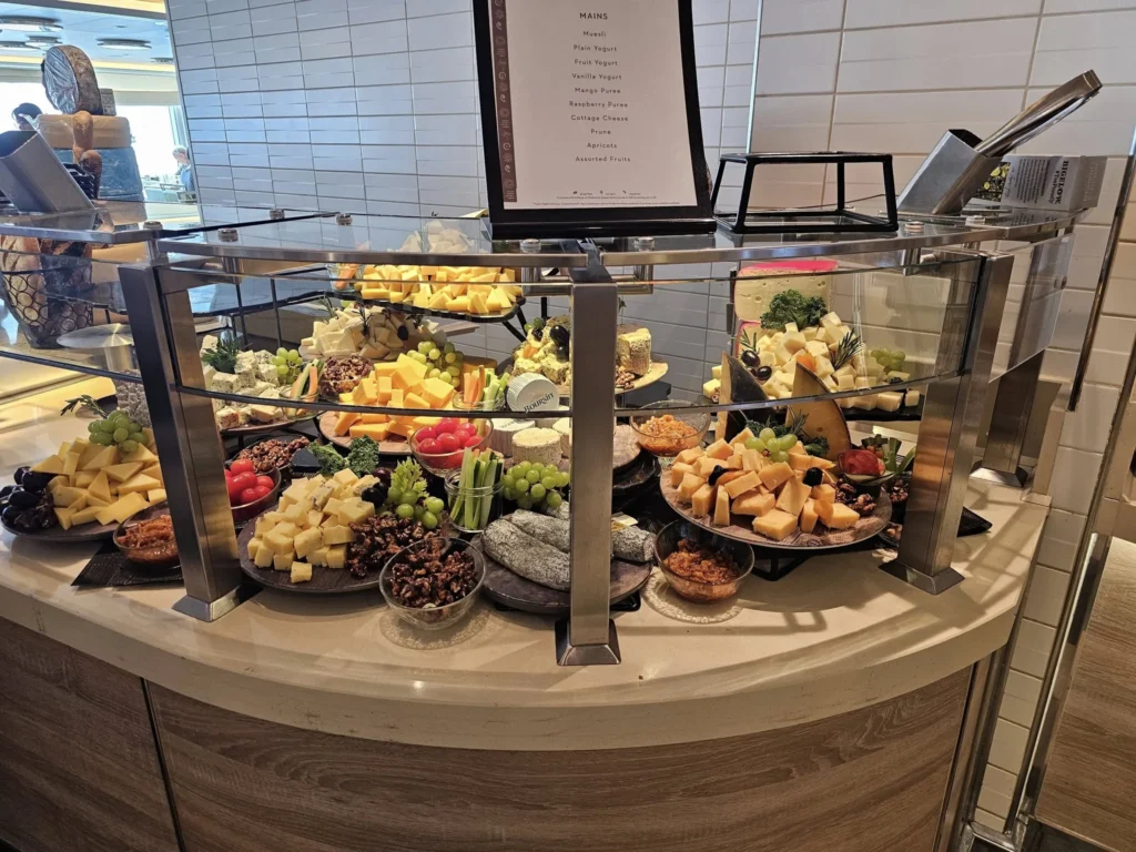 Cruising with Danny and Peter, Holland America Line, Nieuw Statendam, Atlantic Ocean Enchantment Journey Sea Day | A composite image capturing the essence of a luxurious sea day aboard Holland America Line’s Nieuw Statendam cruising the Atlantic Ocean. The scene includes a panoramic view of the vast, shimmering blue Atlantic under a clear sky, with gentle waves and distant seabirds soaring. The elegant interior of the Nieuw Statendam’s Lido Market buffet is shown, featuring a vibrant display of chocolates and cheeses on a table, surrounded by modern decor and large windows framing the ocean. A cozy, warmly lit dining setup at Canaletto, the ship’s Italian specialty restaurant, showcases a beautifully plated osso buco dish with saffron risotto, accompanied by a glass of Chianti. The ship’s sleek exterior glides through the water, its white hull contrasting the deep ocean, with the Mid-Atlantic Ridge subtly illustrated beneath the surface to hint at the ocean’s geological history. A group of passengers enjoys a lively trivia game in the Ocean Bar, with a mysterious camera crew discreetly filming in the background, adding an air of intrigue. The final touch includes a glimpse of a plush stateroom with a large window revealing the moonlit Atlantic, evoking tranquility and anticipation for Agadir, Morocco.