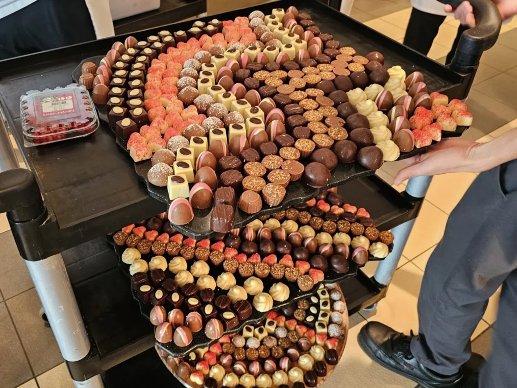 Cruising with Danny and Peter, Holland America Line, Nieuw Statendam, Atlantic Ocean Enchantment Journey Sea Day | A composite image capturing the essence of a luxurious sea day aboard Holland America Line’s Nieuw Statendam cruising the Atlantic Ocean. The scene includes a panoramic view of the vast, shimmering blue Atlantic under a clear sky, with gentle waves and distant seabirds soaring. The elegant interior of the Nieuw Statendam’s Lido Market buffet is shown, featuring a vibrant display of chocolates and cheeses on a table, surrounded by modern decor and large windows framing the ocean. A cozy, warmly lit dining setup at Canaletto, the ship’s Italian specialty restaurant, showcases a beautifully plated osso buco dish with saffron risotto, accompanied by a glass of Chianti. The ship’s sleek exterior glides through the water, its white hull contrasting the deep ocean, with the Mid-Atlantic Ridge subtly illustrated beneath the surface to hint at the ocean’s geological history. A group of passengers enjoys a lively trivia game in the Ocean Bar, with a mysterious camera crew discreetly filming in the background, adding an air of intrigue. The final touch includes a glimpse of a plush stateroom with a large window revealing the moonlit Atlantic, evoking tranquility and anticipation for Agadir, Morocco.