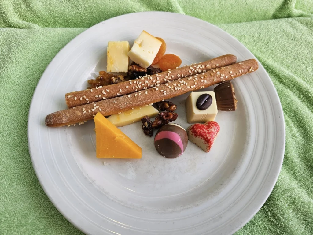 Cruising with Danny and Peter, Holland America Line, Nieuw Statendam, Atlantic Ocean Enchantment Journey Sea Day | A composite image capturing the essence of a luxurious sea day aboard Holland America Line’s Nieuw Statendam cruising the Atlantic Ocean. The scene includes a panoramic view of the vast, shimmering blue Atlantic under a clear sky, with gentle waves and distant seabirds soaring. The elegant interior of the Nieuw Statendam’s Lido Market buffet is shown, featuring a vibrant display of chocolates and cheeses on a table, surrounded by modern decor and large windows framing the ocean. A cozy, warmly lit dining setup at Canaletto, the ship’s Italian specialty restaurant, showcases a beautifully plated osso buco dish with saffron risotto, accompanied by a glass of Chianti. The ship’s sleek exterior glides through the water, its white hull contrasting the deep ocean, with the Mid-Atlantic Ridge subtly illustrated beneath the surface to hint at the ocean’s geological history. A group of passengers enjoys a lively trivia game in the Ocean Bar, with a mysterious camera crew discreetly filming in the background, adding an air of intrigue. The final touch includes a glimpse of a plush stateroom with a large window revealing the moonlit Atlantic, evoking tranquility and anticipation for Agadir, Morocco.