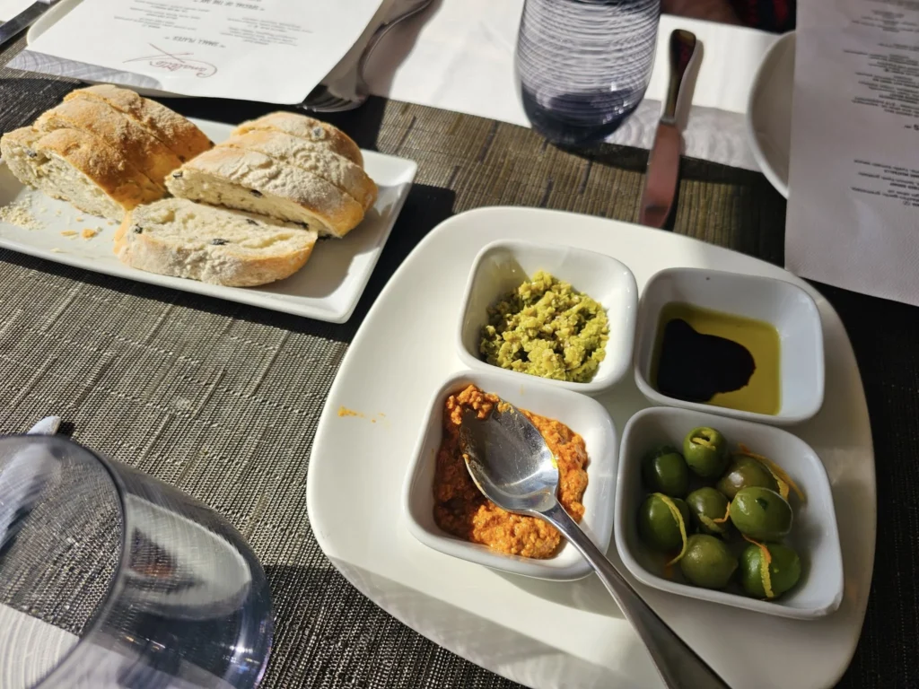 Cruising with Danny and Peter, Holland America Line, Nieuw Statendam, Atlantic Ocean Enchantment Journey Sea Day | A composite image capturing the essence of a luxurious sea day aboard Holland America Line’s Nieuw Statendam cruising the Atlantic Ocean. The scene includes a panoramic view of the vast, shimmering blue Atlantic under a clear sky, with gentle waves and distant seabirds soaring. The elegant interior of the Nieuw Statendam’s Lido Market buffet is shown, featuring a vibrant display of chocolates and cheeses on a table, surrounded by modern decor and large windows framing the ocean. A cozy, warmly lit dining setup at Canaletto, the ship’s Italian specialty restaurant, showcases a beautifully plated osso buco dish with saffron risotto, accompanied by a glass of Chianti. The ship’s sleek exterior glides through the water, its white hull contrasting the deep ocean, with the Mid-Atlantic Ridge subtly illustrated beneath the surface to hint at the ocean’s geological history. A group of passengers enjoys a lively trivia game in the Ocean Bar, with a mysterious camera crew discreetly filming in the background, adding an air of intrigue. The final touch includes a glimpse of a plush stateroom with a large window revealing the moonlit Atlantic, evoking tranquility and anticipation for Agadir, Morocco.