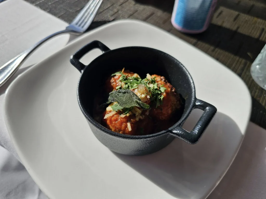 Cruising with Danny and Peter, Holland America Line, Nieuw Statendam, Atlantic Ocean Enchantment Journey Sea Day | A composite image capturing the essence of a luxurious sea day aboard Holland America Line’s Nieuw Statendam cruising the Atlantic Ocean. The scene includes a panoramic view of the vast, shimmering blue Atlantic under a clear sky, with gentle waves and distant seabirds soaring. The elegant interior of the Nieuw Statendam’s Lido Market buffet is shown, featuring a vibrant display of chocolates and cheeses on a table, surrounded by modern decor and large windows framing the ocean. A cozy, warmly lit dining setup at Canaletto, the ship’s Italian specialty restaurant, showcases a beautifully plated osso buco dish with saffron risotto, accompanied by a glass of Chianti. The ship’s sleek exterior glides through the water, its white hull contrasting the deep ocean, with the Mid-Atlantic Ridge subtly illustrated beneath the surface to hint at the ocean’s geological history. A group of passengers enjoys a lively trivia game in the Ocean Bar, with a mysterious camera crew discreetly filming in the background, adding an air of intrigue. The final touch includes a glimpse of a plush stateroom with a large window revealing the moonlit Atlantic, evoking tranquility and anticipation for Agadir, Morocco.