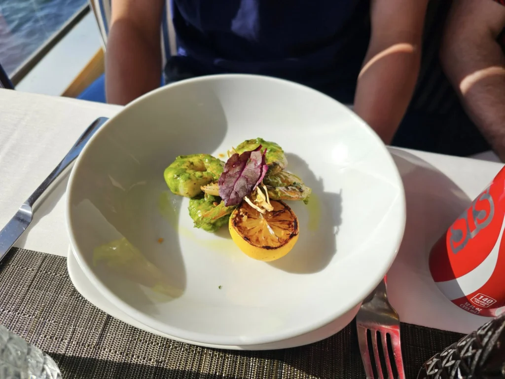 Cruising with Danny and Peter, Holland America Line, Nieuw Statendam, Atlantic Ocean Enchantment Journey Sea Day | A composite image capturing the essence of a luxurious sea day aboard Holland America Line’s Nieuw Statendam cruising the Atlantic Ocean. The scene includes a panoramic view of the vast, shimmering blue Atlantic under a clear sky, with gentle waves and distant seabirds soaring. The elegant interior of the Nieuw Statendam’s Lido Market buffet is shown, featuring a vibrant display of chocolates and cheeses on a table, surrounded by modern decor and large windows framing the ocean. A cozy, warmly lit dining setup at Canaletto, the ship’s Italian specialty restaurant, showcases a beautifully plated osso buco dish with saffron risotto, accompanied by a glass of Chianti. The ship’s sleek exterior glides through the water, its white hull contrasting the deep ocean, with the Mid-Atlantic Ridge subtly illustrated beneath the surface to hint at the ocean’s geological history. A group of passengers enjoys a lively trivia game in the Ocean Bar, with a mysterious camera crew discreetly filming in the background, adding an air of intrigue. The final touch includes a glimpse of a plush stateroom with a large window revealing the moonlit Atlantic, evoking tranquility and anticipation for Agadir, Morocco.