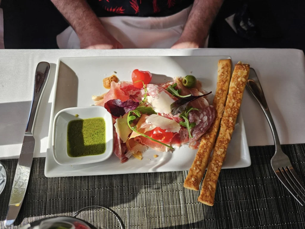 Cruising with Danny and Peter, Holland America Line, Nieuw Statendam, Atlantic Ocean Enchantment Journey Sea Day | A composite image capturing the essence of a luxurious sea day aboard Holland America Line’s Nieuw Statendam cruising the Atlantic Ocean. The scene includes a panoramic view of the vast, shimmering blue Atlantic under a clear sky, with gentle waves and distant seabirds soaring. The elegant interior of the Nieuw Statendam’s Lido Market buffet is shown, featuring a vibrant display of chocolates and cheeses on a table, surrounded by modern decor and large windows framing the ocean. A cozy, warmly lit dining setup at Canaletto, the ship’s Italian specialty restaurant, showcases a beautifully plated osso buco dish with saffron risotto, accompanied by a glass of Chianti. The ship’s sleek exterior glides through the water, its white hull contrasting the deep ocean, with the Mid-Atlantic Ridge subtly illustrated beneath the surface to hint at the ocean’s geological history. A group of passengers enjoys a lively trivia game in the Ocean Bar, with a mysterious camera crew discreetly filming in the background, adding an air of intrigue. The final touch includes a glimpse of a plush stateroom with a large window revealing the moonlit Atlantic, evoking tranquility and anticipation for Agadir, Morocco.