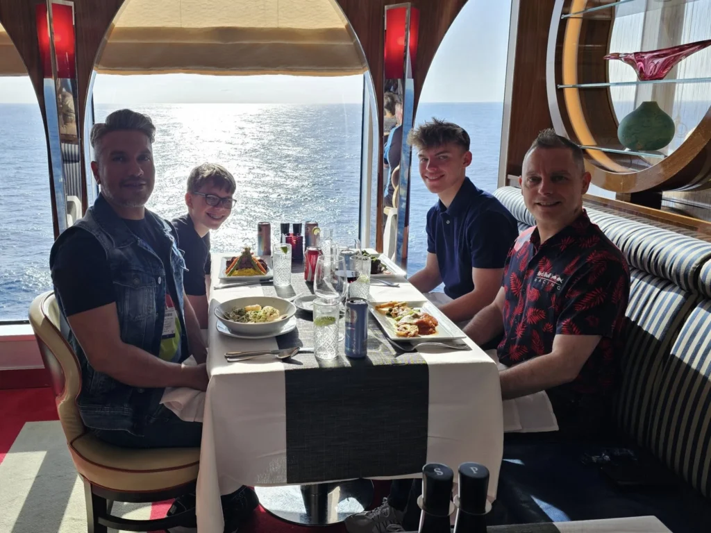 Cruising with Danny and Peter, Holland America Line, Nieuw Statendam, Atlantic Ocean Enchantment Journey Sea Day | A composite image capturing the essence of a luxurious sea day aboard Holland America Line’s Nieuw Statendam cruising the Atlantic Ocean. The scene includes a panoramic view of the vast, shimmering blue Atlantic under a clear sky, with gentle waves and distant seabirds soaring. The elegant interior of the Nieuw Statendam’s Lido Market buffet is shown, featuring a vibrant display of chocolates and cheeses on a table, surrounded by modern decor and large windows framing the ocean. A cozy, warmly lit dining setup at Canaletto, the ship’s Italian specialty restaurant, showcases a beautifully plated osso buco dish with saffron risotto, accompanied by a glass of Chianti. The ship’s sleek exterior glides through the water, its white hull contrasting the deep ocean, with the Mid-Atlantic Ridge subtly illustrated beneath the surface to hint at the ocean’s geological history. A group of passengers enjoys a lively trivia game in the Ocean Bar, with a mysterious camera crew discreetly filming in the background, adding an air of intrigue. The final touch includes a glimpse of a plush stateroom with a large window revealing the moonlit Atlantic, evoking tranquility and anticipation for Agadir, Morocco.