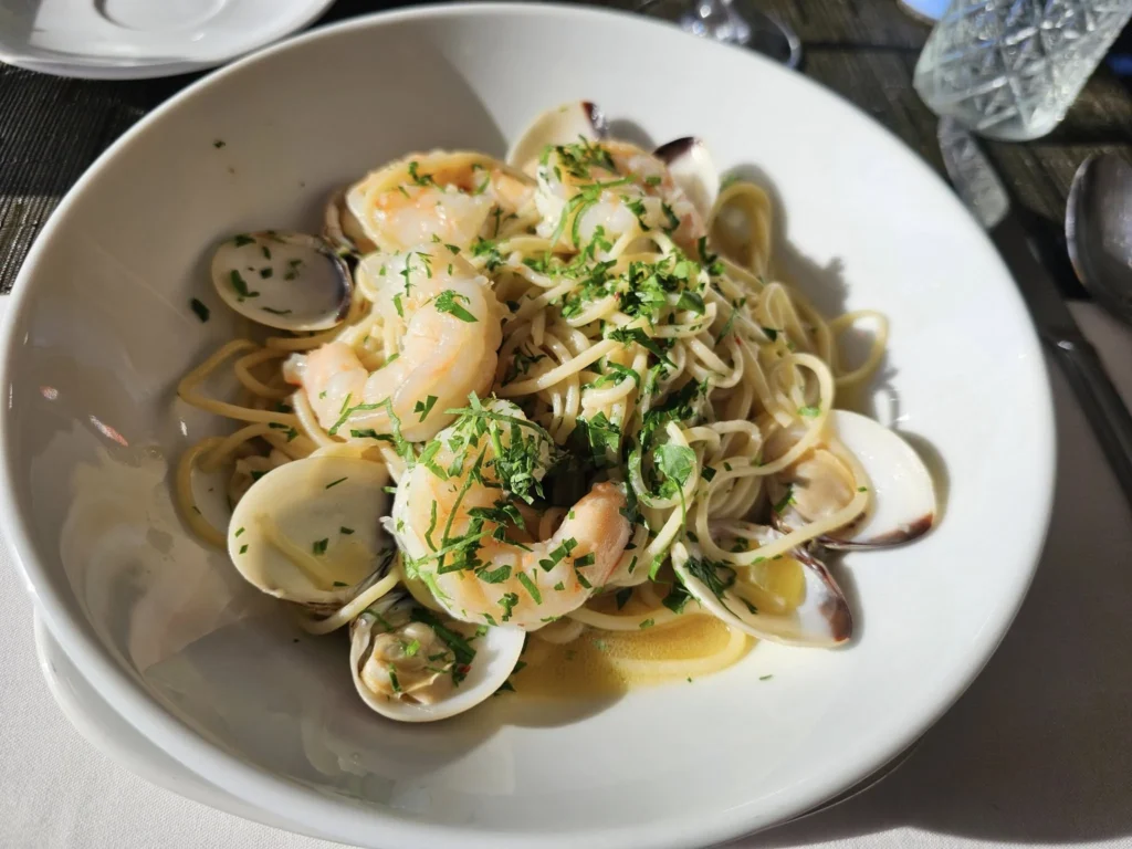 Cruising with Danny and Peter, Holland America Line, Nieuw Statendam, Atlantic Ocean Enchantment Journey Sea Day | A composite image capturing the essence of a luxurious sea day aboard Holland America Line’s Nieuw Statendam cruising the Atlantic Ocean. The scene includes a panoramic view of the vast, shimmering blue Atlantic under a clear sky, with gentle waves and distant seabirds soaring. The elegant interior of the Nieuw Statendam’s Lido Market buffet is shown, featuring a vibrant display of chocolates and cheeses on a table, surrounded by modern decor and large windows framing the ocean. A cozy, warmly lit dining setup at Canaletto, the ship’s Italian specialty restaurant, showcases a beautifully plated osso buco dish with saffron risotto, accompanied by a glass of Chianti. The ship’s sleek exterior glides through the water, its white hull contrasting the deep ocean, with the Mid-Atlantic Ridge subtly illustrated beneath the surface to hint at the ocean’s geological history. A group of passengers enjoys a lively trivia game in the Ocean Bar, with a mysterious camera crew discreetly filming in the background, adding an air of intrigue. The final touch includes a glimpse of a plush stateroom with a large window revealing the moonlit Atlantic, evoking tranquility and anticipation for Agadir, Morocco.