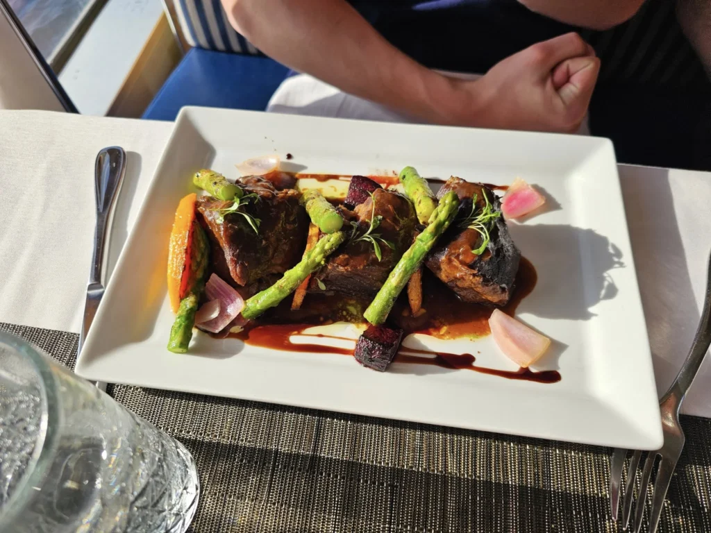 Cruising with Danny and Peter, Holland America Line, Nieuw Statendam, Atlantic Ocean Enchantment Journey Sea Day | A composite image capturing the essence of a luxurious sea day aboard Holland America Line’s Nieuw Statendam cruising the Atlantic Ocean. The scene includes a panoramic view of the vast, shimmering blue Atlantic under a clear sky, with gentle waves and distant seabirds soaring. The elegant interior of the Nieuw Statendam’s Lido Market buffet is shown, featuring a vibrant display of chocolates and cheeses on a table, surrounded by modern decor and large windows framing the ocean. A cozy, warmly lit dining setup at Canaletto, the ship’s Italian specialty restaurant, showcases a beautifully plated osso buco dish with saffron risotto, accompanied by a glass of Chianti. The ship’s sleek exterior glides through the water, its white hull contrasting the deep ocean, with the Mid-Atlantic Ridge subtly illustrated beneath the surface to hint at the ocean’s geological history. A group of passengers enjoys a lively trivia game in the Ocean Bar, with a mysterious camera crew discreetly filming in the background, adding an air of intrigue. The final touch includes a glimpse of a plush stateroom with a large window revealing the moonlit Atlantic, evoking tranquility and anticipation for Agadir, Morocco.