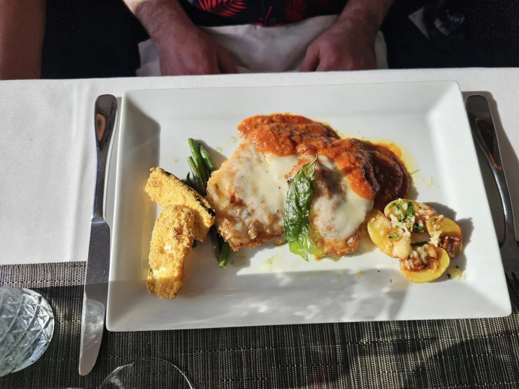 Cruising with Danny and Peter, Holland America Line, Nieuw Statendam, Atlantic Ocean Enchantment Journey Sea Day | A composite image capturing the essence of a luxurious sea day aboard Holland America Line’s Nieuw Statendam cruising the Atlantic Ocean. The scene includes a panoramic view of the vast, shimmering blue Atlantic under a clear sky, with gentle waves and distant seabirds soaring. The elegant interior of the Nieuw Statendam’s Lido Market buffet is shown, featuring a vibrant display of chocolates and cheeses on a table, surrounded by modern decor and large windows framing the ocean. A cozy, warmly lit dining setup at Canaletto, the ship’s Italian specialty restaurant, showcases a beautifully plated osso buco dish with saffron risotto, accompanied by a glass of Chianti. The ship’s sleek exterior glides through the water, its white hull contrasting the deep ocean, with the Mid-Atlantic Ridge subtly illustrated beneath the surface to hint at the ocean’s geological history. A group of passengers enjoys a lively trivia game in the Ocean Bar, with a mysterious camera crew discreetly filming in the background, adding an air of intrigue. The final touch includes a glimpse of a plush stateroom with a large window revealing the moonlit Atlantic, evoking tranquility and anticipation for Agadir, Morocco.