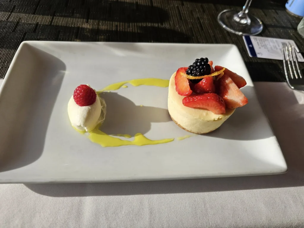 Cruising with Danny and Peter, Holland America Line, Nieuw Statendam, Atlantic Ocean Enchantment Journey Sea Day | A composite image capturing the essence of a luxurious sea day aboard Holland America Line’s Nieuw Statendam cruising the Atlantic Ocean. The scene includes a panoramic view of the vast, shimmering blue Atlantic under a clear sky, with gentle waves and distant seabirds soaring. The elegant interior of the Nieuw Statendam’s Lido Market buffet is shown, featuring a vibrant display of chocolates and cheeses on a table, surrounded by modern decor and large windows framing the ocean. A cozy, warmly lit dining setup at Canaletto, the ship’s Italian specialty restaurant, showcases a beautifully plated osso buco dish with saffron risotto, accompanied by a glass of Chianti. The ship’s sleek exterior glides through the water, its white hull contrasting the deep ocean, with the Mid-Atlantic Ridge subtly illustrated beneath the surface to hint at the ocean’s geological history. A group of passengers enjoys a lively trivia game in the Ocean Bar, with a mysterious camera crew discreetly filming in the background, adding an air of intrigue. The final touch includes a glimpse of a plush stateroom with a large window revealing the moonlit Atlantic, evoking tranquility and anticipation for Agadir, Morocco.