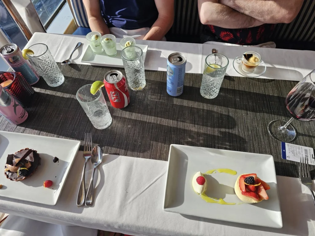 Cruising with Danny and Peter, Holland America Line, Nieuw Statendam, Atlantic Ocean Enchantment Journey Sea Day | A composite image capturing the essence of a luxurious sea day aboard Holland America Line’s Nieuw Statendam cruising the Atlantic Ocean. The scene includes a panoramic view of the vast, shimmering blue Atlantic under a clear sky, with gentle waves and distant seabirds soaring. The elegant interior of the Nieuw Statendam’s Lido Market buffet is shown, featuring a vibrant display of chocolates and cheeses on a table, surrounded by modern decor and large windows framing the ocean. A cozy, warmly lit dining setup at Canaletto, the ship’s Italian specialty restaurant, showcases a beautifully plated osso buco dish with saffron risotto, accompanied by a glass of Chianti. The ship’s sleek exterior glides through the water, its white hull contrasting the deep ocean, with the Mid-Atlantic Ridge subtly illustrated beneath the surface to hint at the ocean’s geological history. A group of passengers enjoys a lively trivia game in the Ocean Bar, with a mysterious camera crew discreetly filming in the background, adding an air of intrigue. The final touch includes a glimpse of a plush stateroom with a large window revealing the moonlit Atlantic, evoking tranquility and anticipation for Agadir, Morocco.