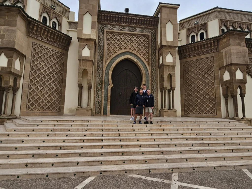 Cruising with Danny and Peter, Holland America Line, Nieuw Statendam, Agadir Morocco Adventure | A vibrant collage capturing a day in Agadir, Morocco, during a Holland America Nieuw Statendam cruise: the white façade and green-tiled minaret of Mosque Mohammed V gleaming under the sun; the bustling Agadir Marina with yachts bobbing in the harbor and golden beach sands stretching toward the Anti-Atlas Mountains; the historic Agadir Oufella kasbah ruins on a hill overlooking the sparkling Atlantic; Sven riding a camel along a scenic path with a Berber handler; the colorful chaos of Souk El Had market with stalls of spices, rugs, and jewelry; the lush Paradise Valley oasis with palm trees, clear pools, and tiny fish nibbling at feet; and an aromatic garden with Moroccan bread, argan oil, and mint tea under a shaded canopy.