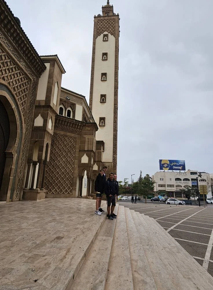 Cruising with Danny and Peter, Holland America Line, Nieuw Statendam, Agadir Morocco Adventure | A vibrant collage capturing a day in Agadir, Morocco, during a Holland America Nieuw Statendam cruise: the white façade and green-tiled minaret of Mosque Mohammed V gleaming under the sun; the bustling Agadir Marina with yachts bobbing in the harbor and golden beach sands stretching toward the Anti-Atlas Mountains; the historic Agadir Oufella kasbah ruins on a hill overlooking the sparkling Atlantic; Sven riding a camel along a scenic path with a Berber handler; the colorful chaos of Souk El Had market with stalls of spices, rugs, and jewelry; the lush Paradise Valley oasis with palm trees, clear pools, and tiny fish nibbling at feet; and an aromatic garden with Moroccan bread, argan oil, and mint tea under a shaded canopy.