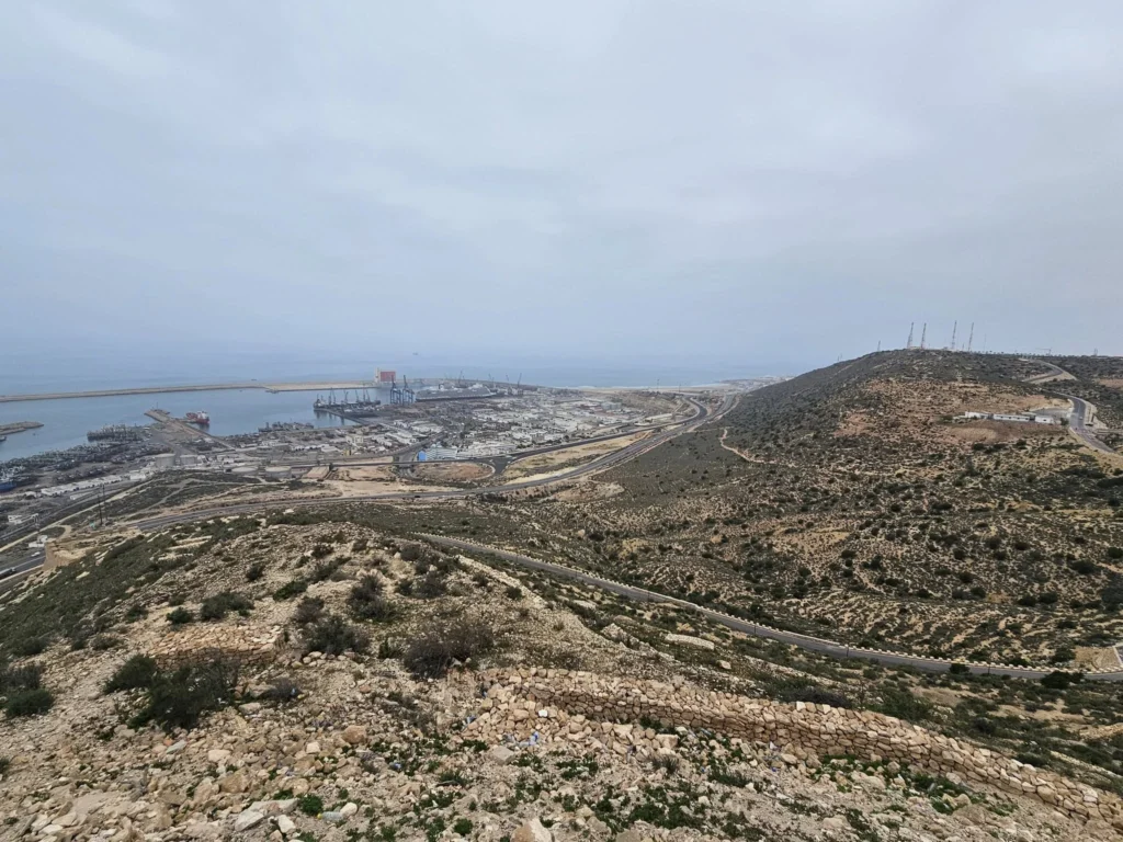 Cruising with Danny and Peter, Holland America Line, Nieuw Statendam, Agadir Morocco Adventure | A vibrant collage capturing a day in Agadir, Morocco, during a Holland America Nieuw Statendam cruise: the white façade and green-tiled minaret of Mosque Mohammed V gleaming under the sun; the bustling Agadir Marina with yachts bobbing in the harbor and golden beach sands stretching toward the Anti-Atlas Mountains; the historic Agadir Oufella kasbah ruins on a hill overlooking the sparkling Atlantic; Sven riding a camel along a scenic path with a Berber handler; the colorful chaos of Souk El Had market with stalls of spices, rugs, and jewelry; the lush Paradise Valley oasis with palm trees, clear pools, and tiny fish nibbling at feet; and an aromatic garden with Moroccan bread, argan oil, and mint tea under a shaded canopy.