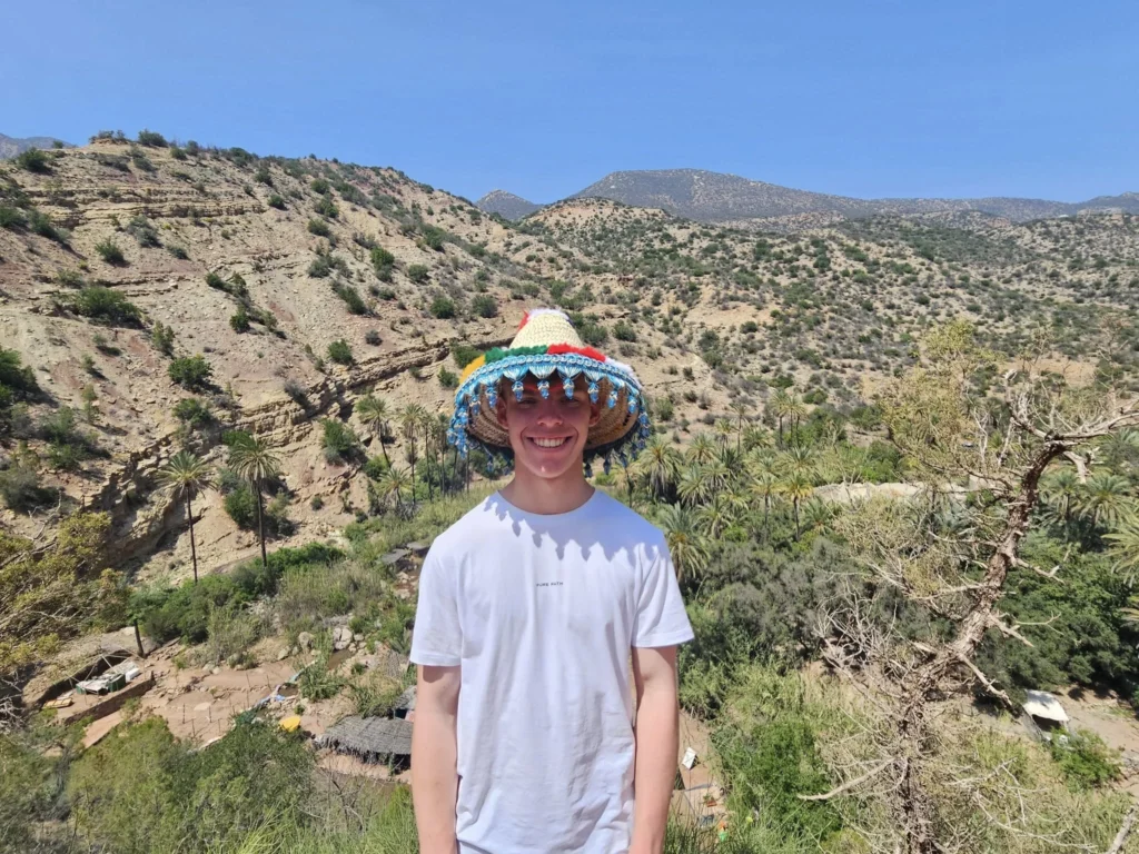 Cruising with Danny and Peter, Holland America Line, Nieuw Statendam, Agadir Morocco Adventure | A vibrant collage capturing a day in Agadir, Morocco, during a Holland America Nieuw Statendam cruise: the white façade and green-tiled minaret of Mosque Mohammed V gleaming under the sun; the bustling Agadir Marina with yachts bobbing in the harbor and golden beach sands stretching toward the Anti-Atlas Mountains; the historic Agadir Oufella kasbah ruins on a hill overlooking the sparkling Atlantic; Sven riding a camel along a scenic path with a Berber handler; the colorful chaos of Souk El Had market with stalls of spices, rugs, and jewelry; the lush Paradise Valley oasis with palm trees, clear pools, and tiny fish nibbling at feet; and an aromatic garden with Moroccan bread, argan oil, and mint tea under a shaded canopy.