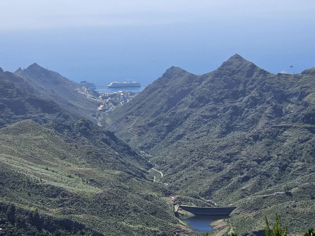 Cruising with Danny and Peter, Holland America Line, Nieuw Statendam, Santa Cruz, Tenerife, Canary Islands, History Adventure | A vibrant collage capturing Santa Cruz, Tenerife’s diverse beauty: the misty, lush laurel forests of Anaga Rural Park with sunlight filtering through dense green foliage at Mirador Cruz del Carmen; a panoramic view from Mirador Pico del Inglés showcasing rugged Anaga Mountains and the distant Atlantic Ocean under a partly cloudy sky; the golden sands of Playa de las Teresitas lined with palm trees and calm turquoise waters; a colorful seafood platter with grilled prawns and papas arrugadas served at a beachfront restaurant; and the elegant Nieuw Statendam cruise ship docked at Santa Cruz port with the city’s skyline and Anaga Mountains in the background.
