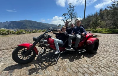Cruising with Danny and Peter, Holland America Line, Nieuw Statendam, Funchal, Madeira. Portugal, History Adventure | A vibrant collage capturing a day in Funchal, Madeira: a sleek trike navigates winding roads with lush green hills in the background; the panoramic view from Pico dos Barcelos showcases Funchal Bay and the Desertas Islands under a clear sky; Cascata dos Anjos waterfall cascades from cliffs onto a road, surrounded by verdant greenery; Praia da Madalena do Mar’s pebbled beach and black volcanic sand meet gentle waves with towering cliffs above; the misty Valley of the Nuns nestles between rugged mountains, dotted with stone houses and chestnut groves; Cabo Girão’s glass-bottomed skywalk reveals a dizzying drop to the Atlantic and terraced fields below; a plate of warm bolo do caco flatbread filled with steak sits on a café table in Funchal’s colorful old town; the CR7 Museum displays Cristiano Ronaldo’s trophies and jerseys with a waterfront view of the Nieuw Statendam; and Nami Sushi Bar’s elegant sushi platter gleams under soft lighting aboard the ship.