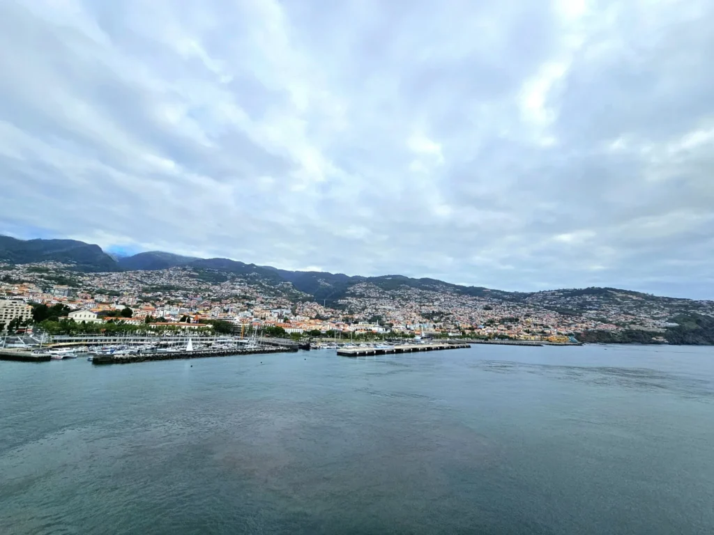 Cruising with Danny and Peter, Holland America Line, Nieuw Statendam, Funchal, Madeira. Portugal, History Adventure | A vibrant collage capturing a day in Funchal, Madeira: a sleek trike navigates winding roads with lush green hills in the background; the panoramic view from Pico dos Barcelos showcases Funchal Bay and the Desertas Islands under a clear sky; Cascata dos Anjos waterfall cascades from cliffs onto a road, surrounded by verdant greenery; Praia da Madalena do Mar’s pebbled beach and black volcanic sand meet gentle waves with towering cliffs above; the misty Valley of the Nuns nestles between rugged mountains, dotted with stone houses and chestnut groves; Cabo Girão’s glass-bottomed skywalk reveals a dizzying drop to the Atlantic and terraced fields below; a plate of warm bolo do caco flatbread filled with steak sits on a café table in Funchal’s colorful old town; the CR7 Museum displays Cristiano Ronaldo’s trophies and jerseys with a waterfront view of the Nieuw Statendam; and Nami Sushi Bar’s elegant sushi platter gleams under soft lighting aboard the ship.