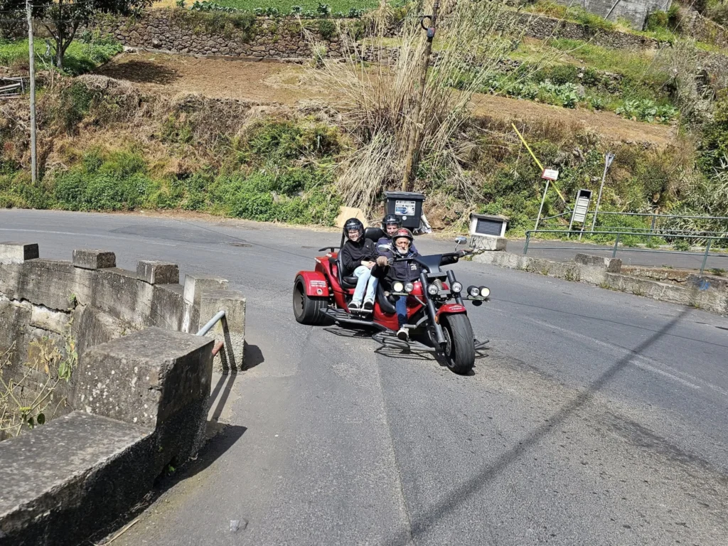 Cruising with Danny and Peter, Holland America Line, Nieuw Statendam, Funchal, Madeira. Portugal, History Adventure | A vibrant collage capturing a day in Funchal, Madeira: a sleek trike navigates winding roads with lush green hills in the background; the panoramic view from Pico dos Barcelos showcases Funchal Bay and the Desertas Islands under a clear sky; Cascata dos Anjos waterfall cascades from cliffs onto a road, surrounded by verdant greenery; Praia da Madalena do Mar’s pebbled beach and black volcanic sand meet gentle waves with towering cliffs above; the misty Valley of the Nuns nestles between rugged mountains, dotted with stone houses and chestnut groves; Cabo Girão’s glass-bottomed skywalk reveals a dizzying drop to the Atlantic and terraced fields below; a plate of warm bolo do caco flatbread filled with steak sits on a café table in Funchal’s colorful old town; the CR7 Museum displays Cristiano Ronaldo’s trophies and jerseys with a waterfront view of the Nieuw Statendam; and Nami Sushi Bar’s elegant sushi platter gleams under soft lighting aboard the ship.