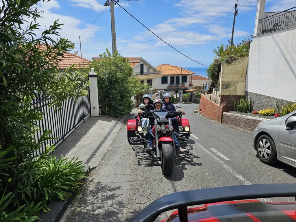 Cruising with Danny and Peter, Holland America Line, Nieuw Statendam, Funchal, Madeira. Portugal, History Adventure | A vibrant collage capturing a day in Funchal, Madeira: a sleek trike navigates winding roads with lush green hills in the background; the panoramic view from Pico dos Barcelos showcases Funchal Bay and the Desertas Islands under a clear sky; Cascata dos Anjos waterfall cascades from cliffs onto a road, surrounded by verdant greenery; Praia da Madalena do Mar’s pebbled beach and black volcanic sand meet gentle waves with towering cliffs above; the misty Valley of the Nuns nestles between rugged mountains, dotted with stone houses and chestnut groves; Cabo Girão’s glass-bottomed skywalk reveals a dizzying drop to the Atlantic and terraced fields below; a plate of warm bolo do caco flatbread filled with steak sits on a café table in Funchal’s colorful old town; the CR7 Museum displays Cristiano Ronaldo’s trophies and jerseys with a waterfront view of the Nieuw Statendam; and Nami Sushi Bar’s elegant sushi platter gleams under soft lighting aboard the ship.
