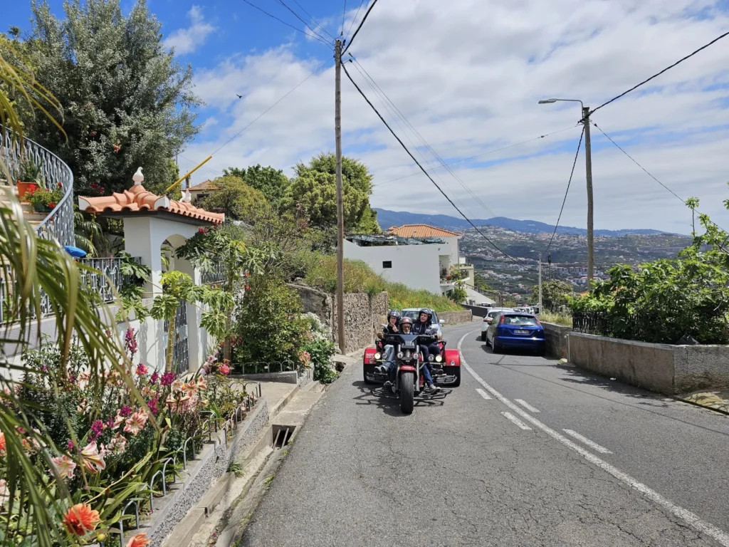 Cruising with Danny and Peter, Holland America Line, Nieuw Statendam, Funchal, Madeira. Portugal, History Adventure | A vibrant collage capturing a day in Funchal, Madeira: a sleek trike navigates winding roads with lush green hills in the background; the panoramic view from Pico dos Barcelos showcases Funchal Bay and the Desertas Islands under a clear sky; Cascata dos Anjos waterfall cascades from cliffs onto a road, surrounded by verdant greenery; Praia da Madalena do Mar’s pebbled beach and black volcanic sand meet gentle waves with towering cliffs above; the misty Valley of the Nuns nestles between rugged mountains, dotted with stone houses and chestnut groves; Cabo Girão’s glass-bottomed skywalk reveals a dizzying drop to the Atlantic and terraced fields below; a plate of warm bolo do caco flatbread filled with steak sits on a café table in Funchal’s colorful old town; the CR7 Museum displays Cristiano Ronaldo’s trophies and jerseys with a waterfront view of the Nieuw Statendam; and Nami Sushi Bar’s elegant sushi platter gleams under soft lighting aboard the ship.