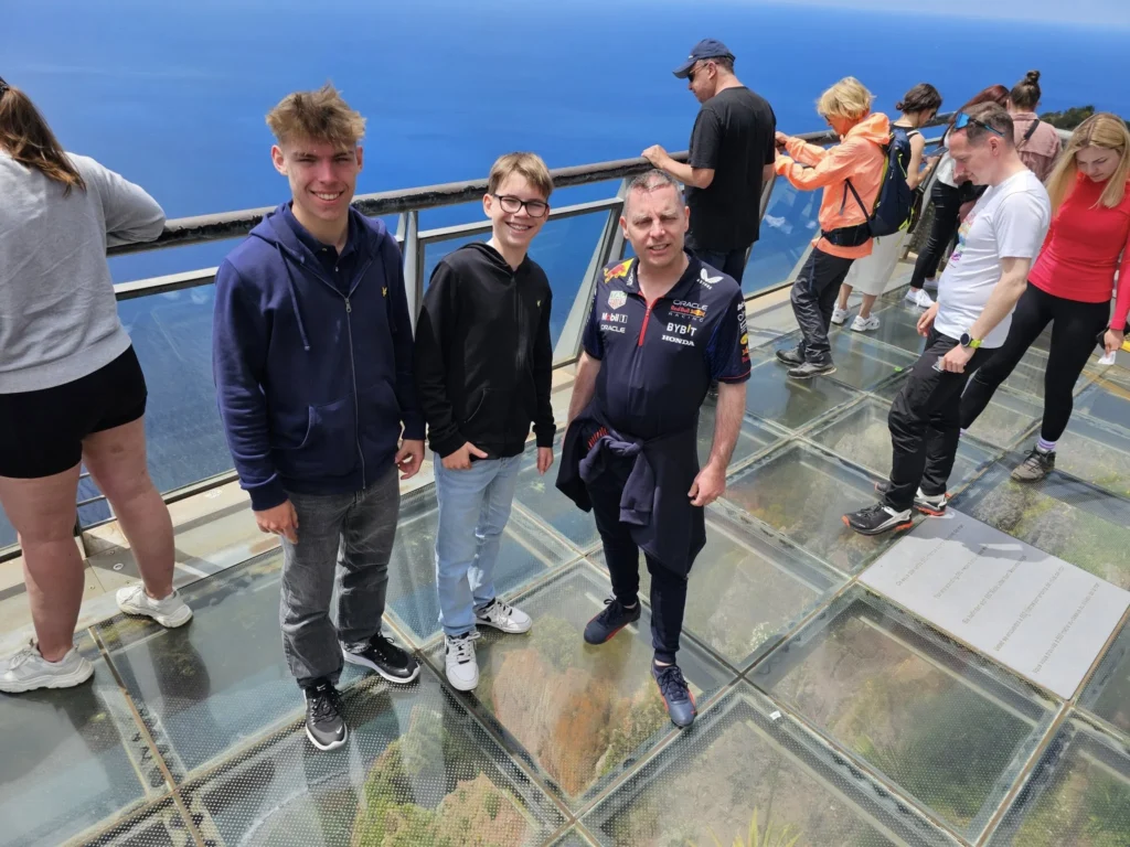 Cruising with Danny and Peter, Holland America Line, Nieuw Statendam, Funchal, Madeira. Portugal, History Adventure | A vibrant collage capturing a day in Funchal, Madeira: a sleek trike navigates winding roads with lush green hills in the background; the panoramic view from Pico dos Barcelos showcases Funchal Bay and the Desertas Islands under a clear sky; Cascata dos Anjos waterfall cascades from cliffs onto a road, surrounded by verdant greenery; Praia da Madalena do Mar’s pebbled beach and black volcanic sand meet gentle waves with towering cliffs above; the misty Valley of the Nuns nestles between rugged mountains, dotted with stone houses and chestnut groves; Cabo Girão’s glass-bottomed skywalk reveals a dizzying drop to the Atlantic and terraced fields below; a plate of warm bolo do caco flatbread filled with steak sits on a café table in Funchal’s colorful old town; the CR7 Museum displays Cristiano Ronaldo’s trophies and jerseys with a waterfront view of the Nieuw Statendam; and Nami Sushi Bar’s elegant sushi platter gleams under soft lighting aboard the ship.
