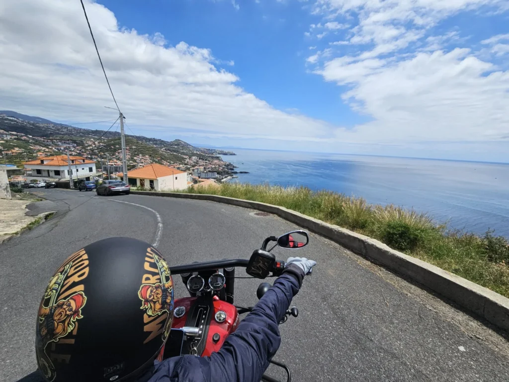 Cruising with Danny and Peter, Holland America Line, Nieuw Statendam, Funchal, Madeira. Portugal, History Adventure | A vibrant collage capturing a day in Funchal, Madeira: a sleek trike navigates winding roads with lush green hills in the background; the panoramic view from Pico dos Barcelos showcases Funchal Bay and the Desertas Islands under a clear sky; Cascata dos Anjos waterfall cascades from cliffs onto a road, surrounded by verdant greenery; Praia da Madalena do Mar’s pebbled beach and black volcanic sand meet gentle waves with towering cliffs above; the misty Valley of the Nuns nestles between rugged mountains, dotted with stone houses and chestnut groves; Cabo Girão’s glass-bottomed skywalk reveals a dizzying drop to the Atlantic and terraced fields below; a plate of warm bolo do caco flatbread filled with steak sits on a café table in Funchal’s colorful old town; the CR7 Museum displays Cristiano Ronaldo’s trophies and jerseys with a waterfront view of the Nieuw Statendam; and Nami Sushi Bar’s elegant sushi platter gleams under soft lighting aboard the ship.